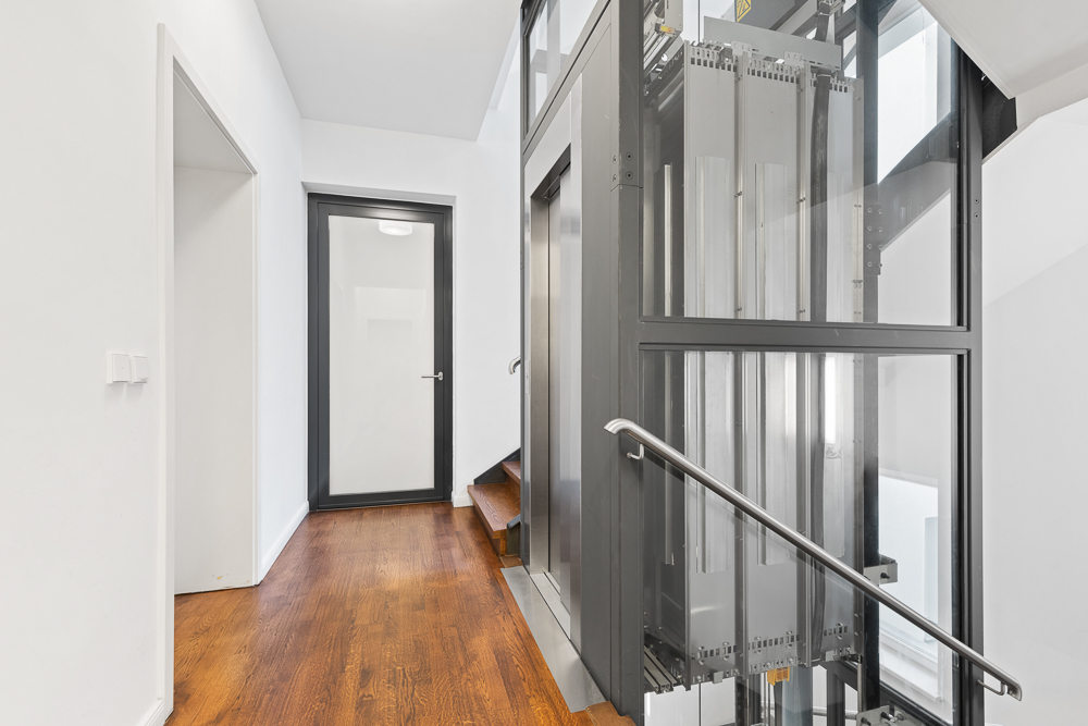 Flur mit Holzboden, Glastür, Aufzug und angrenzender Treppe in einem Wohnhaus.