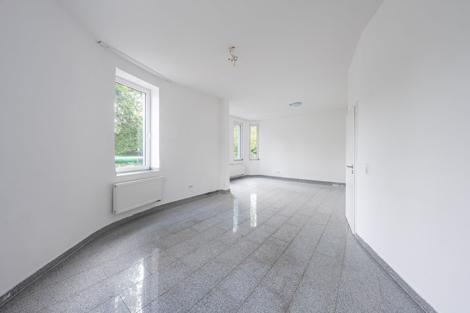 Heller, leerer Raum mit weißen Wänden, grauem Fliesenboden und mehreren Fenstern.