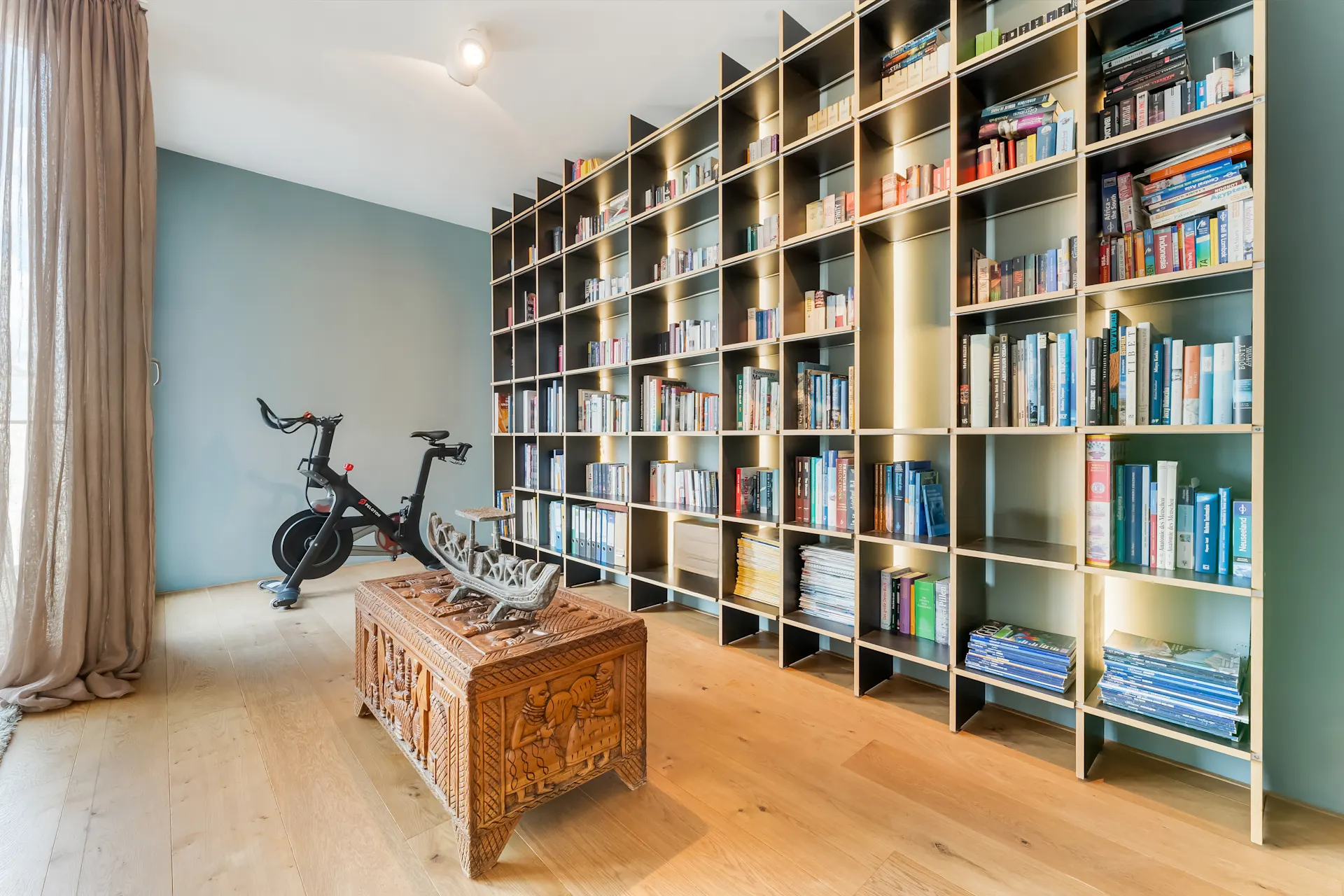 Raum mit großem Bücherregal, Holztisch und einem Heimtrainer auf Holzboden.