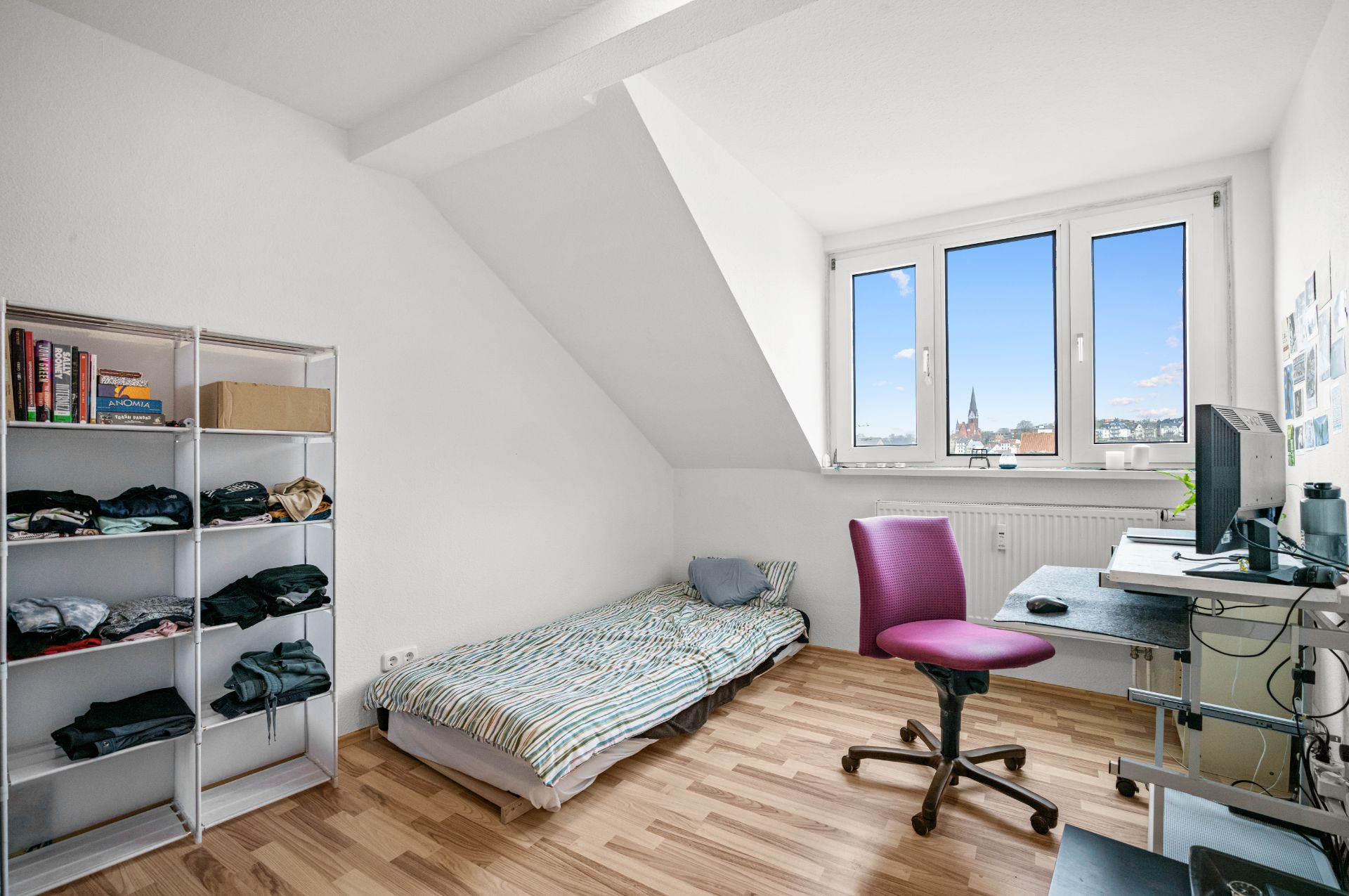 Helles Zimmer mit Einzelbett, Schreibtisch, Stuhl, Regal und großem Fenster mit Ausblick.