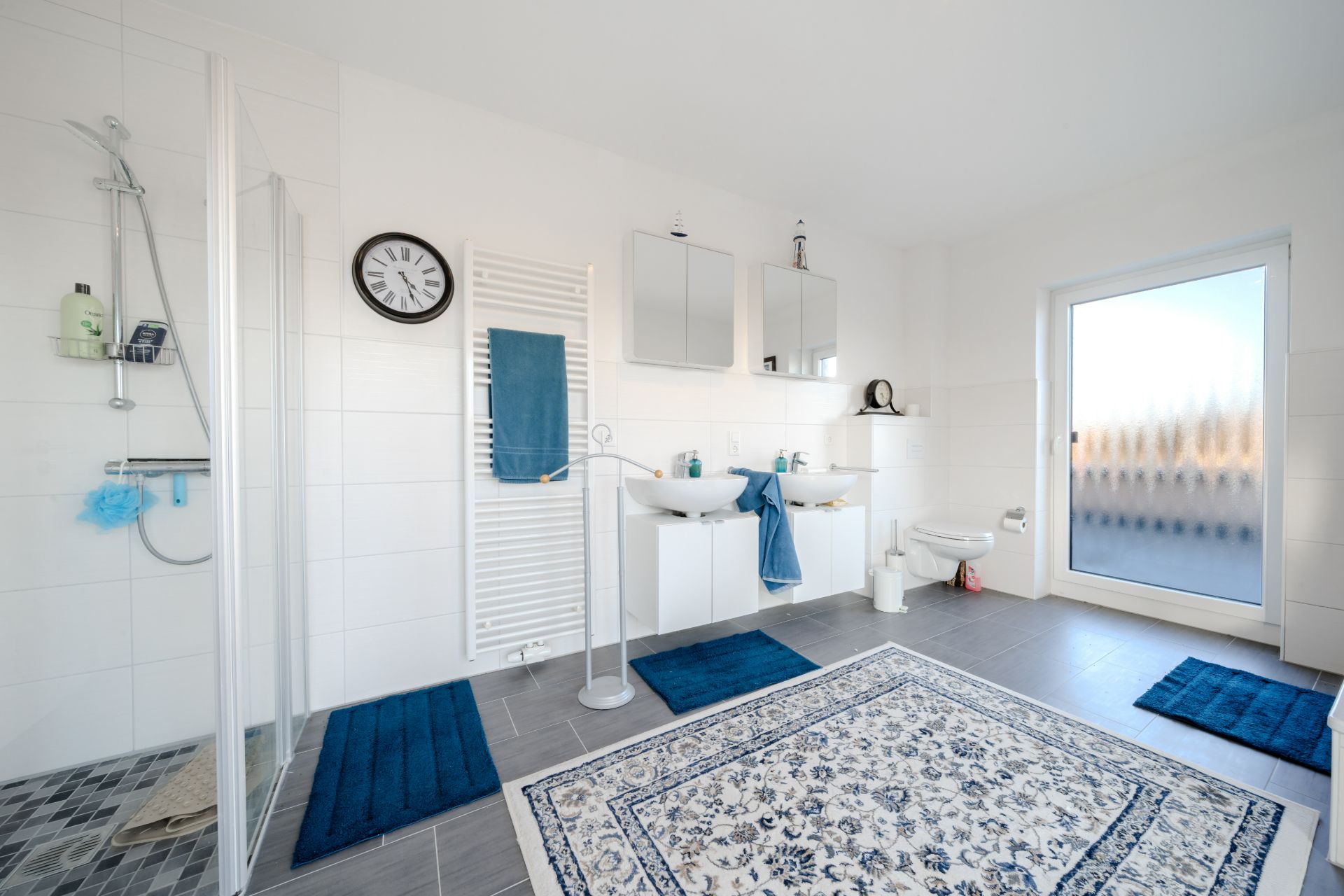 Helles Badezimmer mit Dusche, Doppelwaschbecken, WC und blauen Teppichen.
