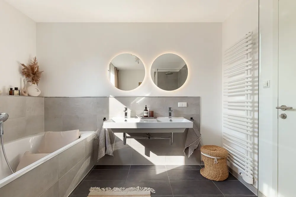 Modernes Badezimmer mit Badewanne, Doppelwaschbecken, zwei runden Spiegeln und Handtuchheizkörper.