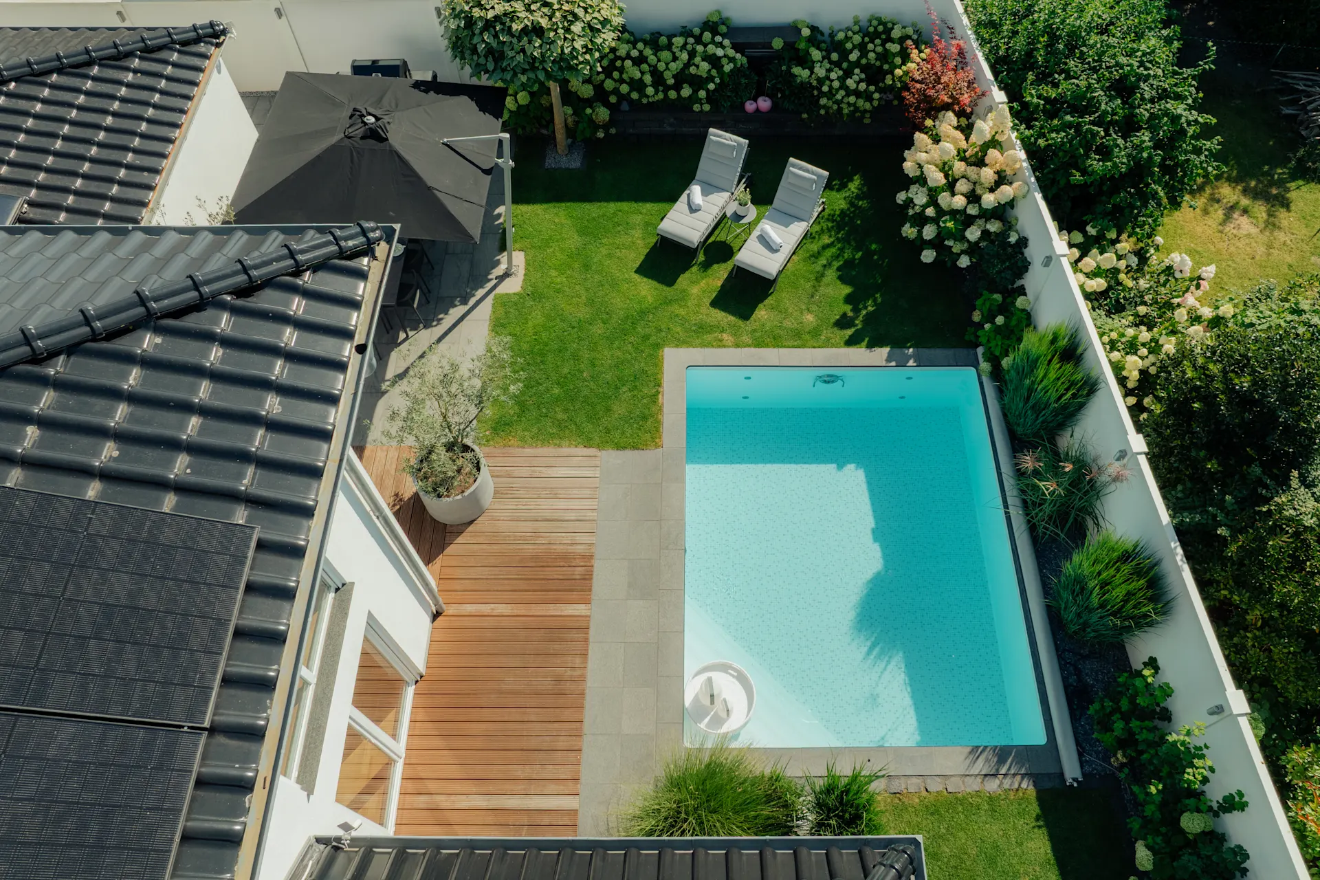 Moderner Garten mit Pool, Holzterrasse, Sonnenschirm, zwei Liegestühlen und Blumenecke.