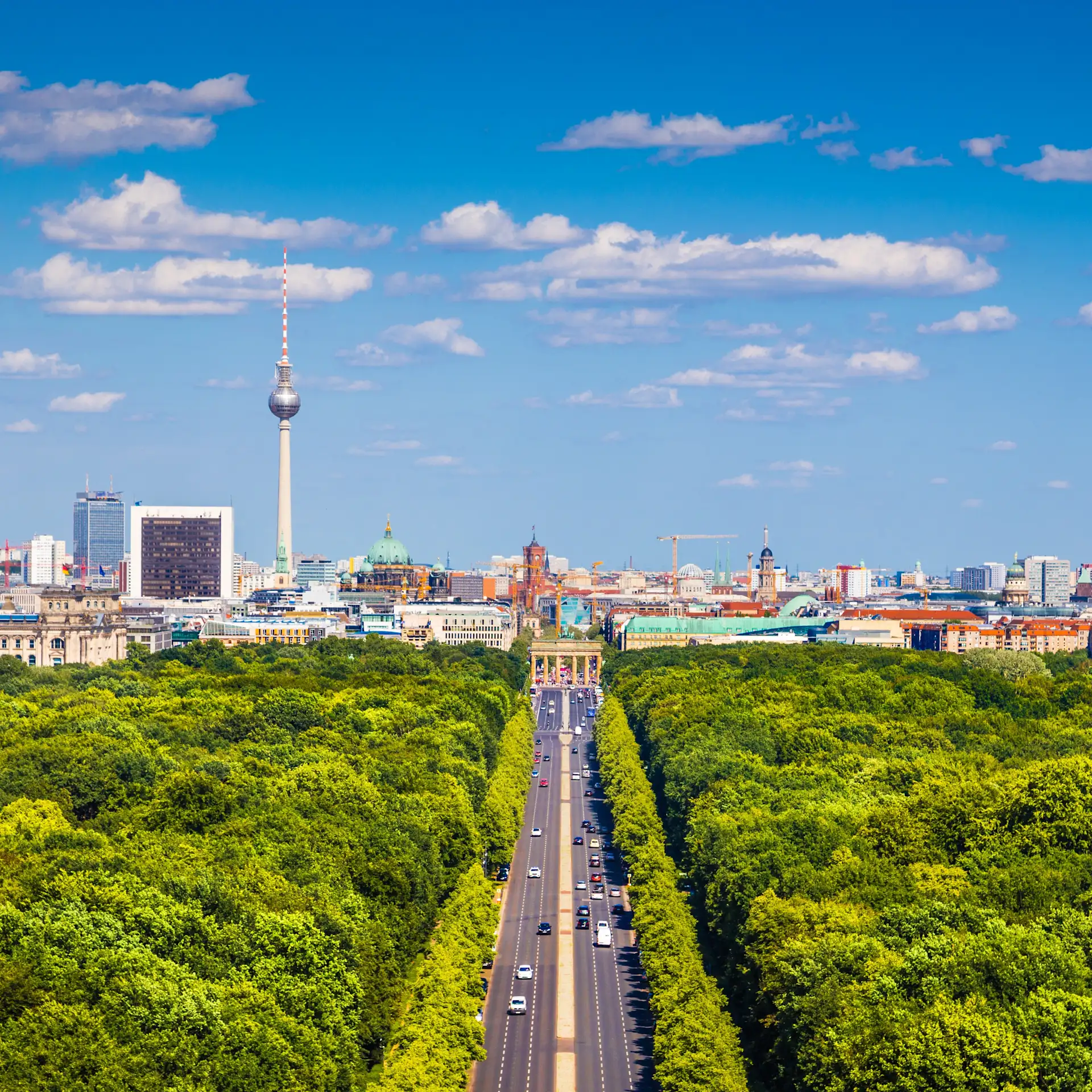 Stadtblick mit breiter Straße durch einen grünen Park und Fernsehturm im Hintergrund.