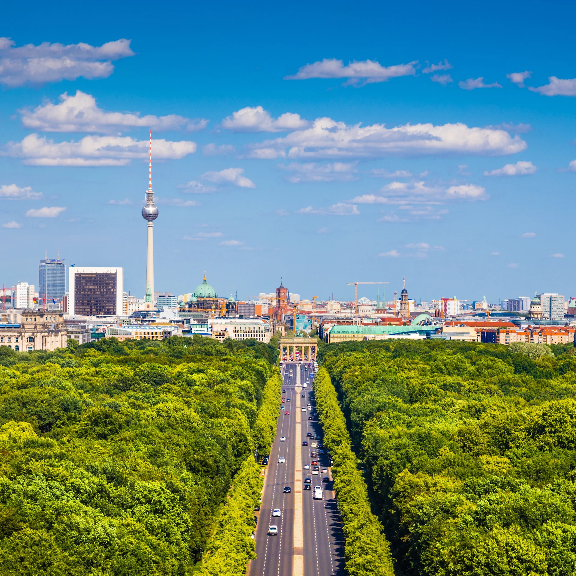 Stadtblick mit breiter Straße durch einen grünen Park und Fernsehturm im Hintergrund.