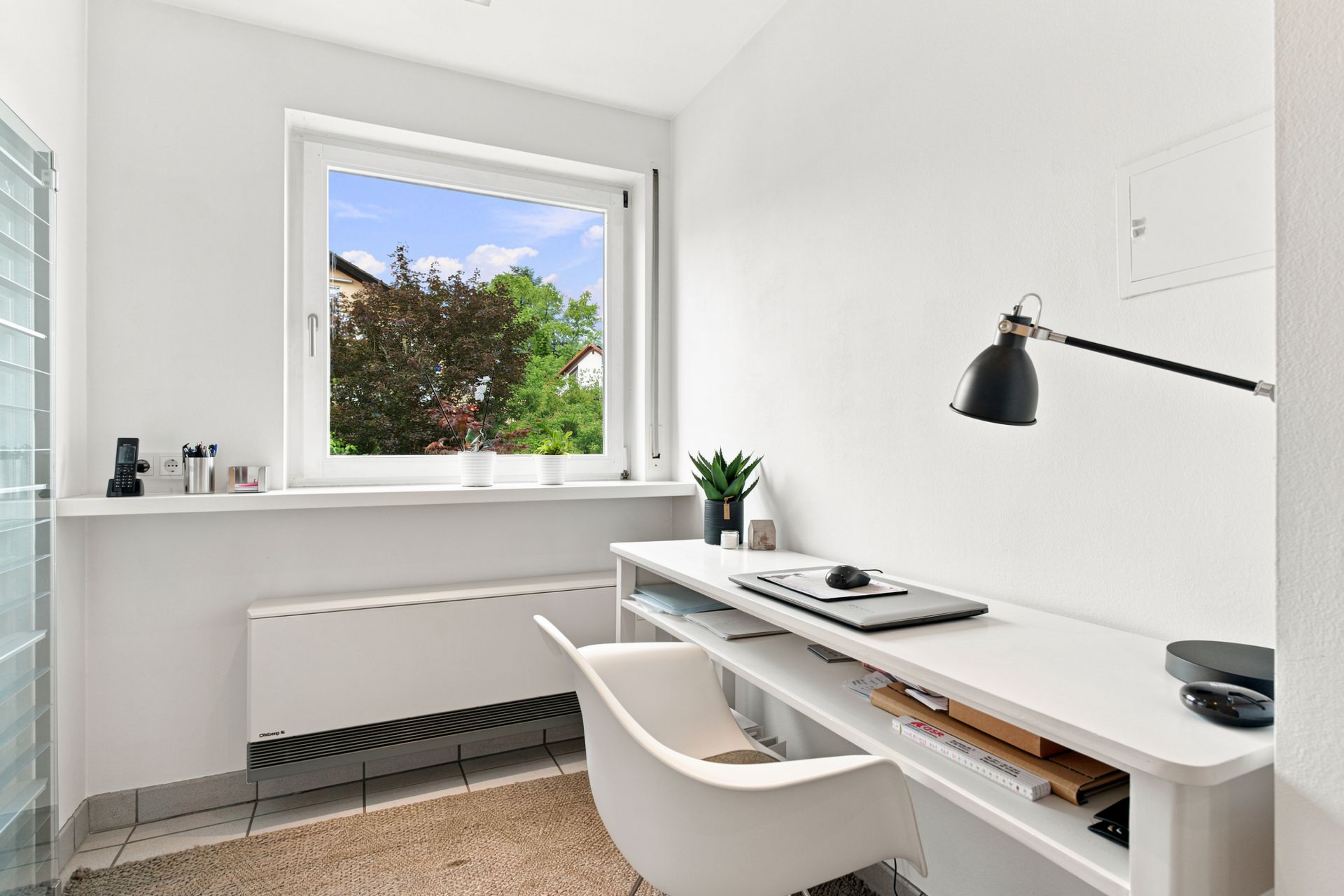 Helles Arbeitszimmer mit Schreibtisch, Stuhl und Lampe vor einem Fenster mit Blick ins Grüne.