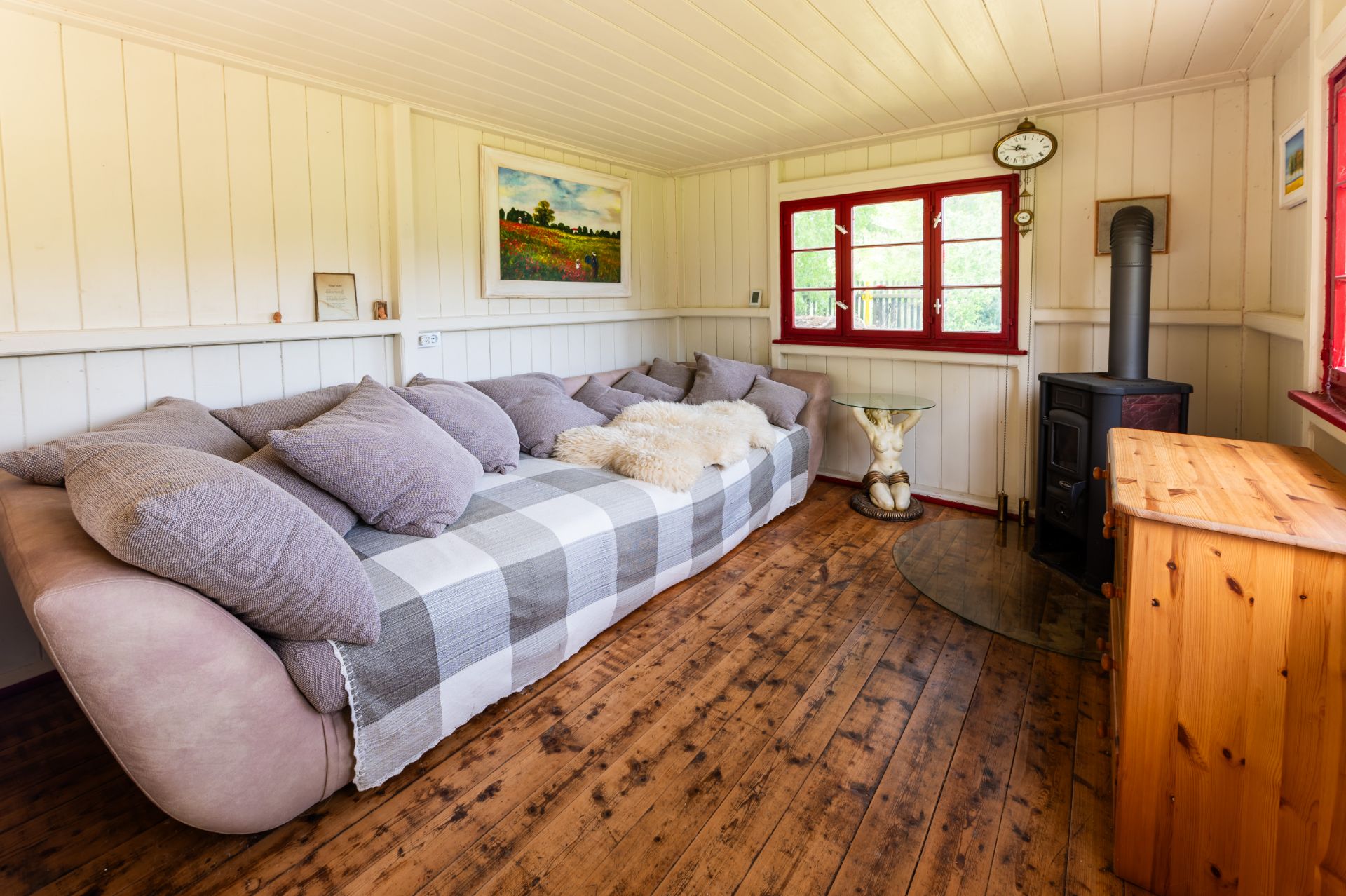 Gemütliches Wohnzimmer mit großem Sofa, Holzboden, Holzofen und roten Fenstern.
