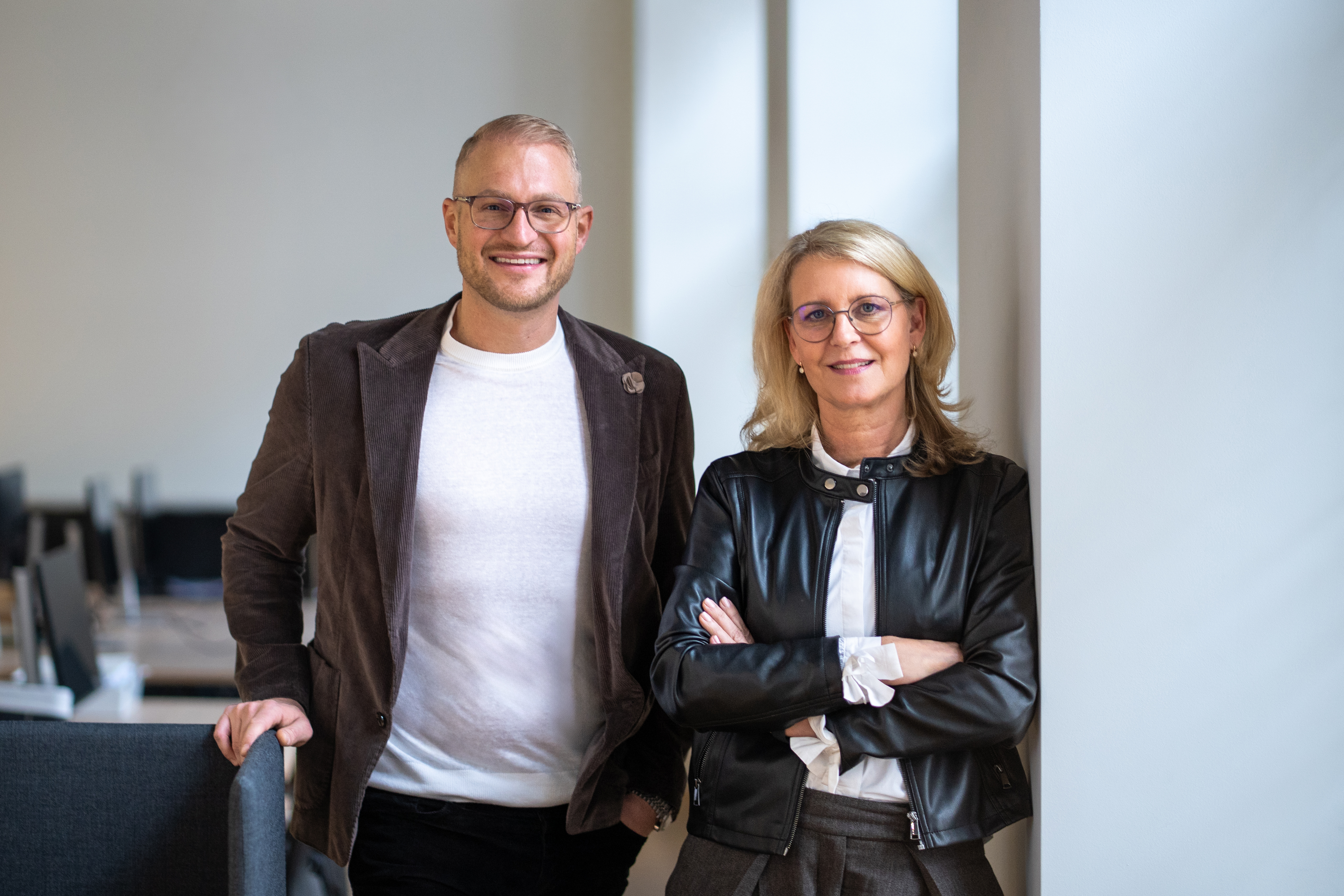 Ein Mann und eine Frau mit Brillen stehen lächelnd in einem modernen Raum mit Tischen.