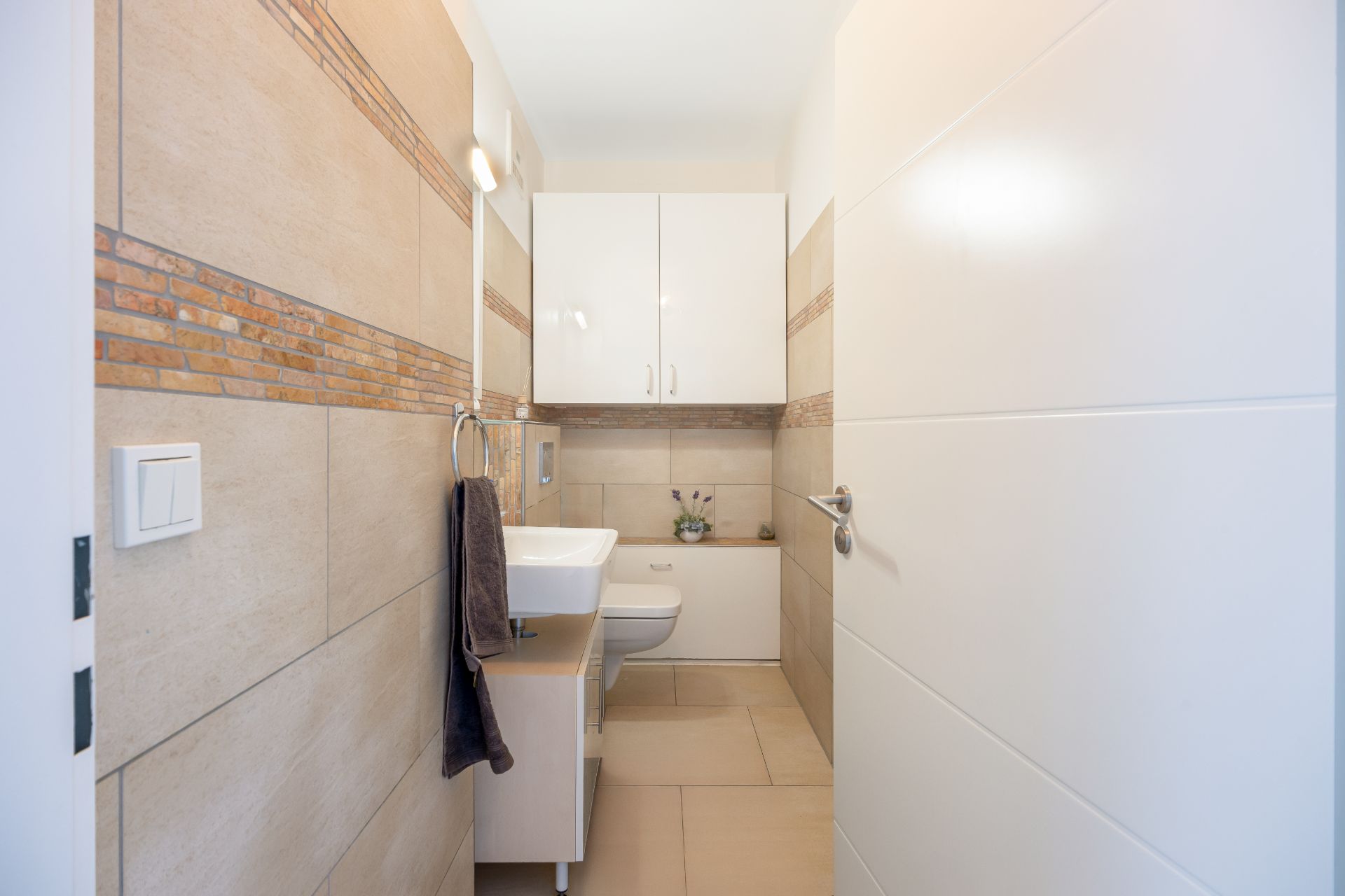 Modernes Badezimmer mit Wandfliesen, Waschbecken, WC, Schrank und Handtuchhalter.