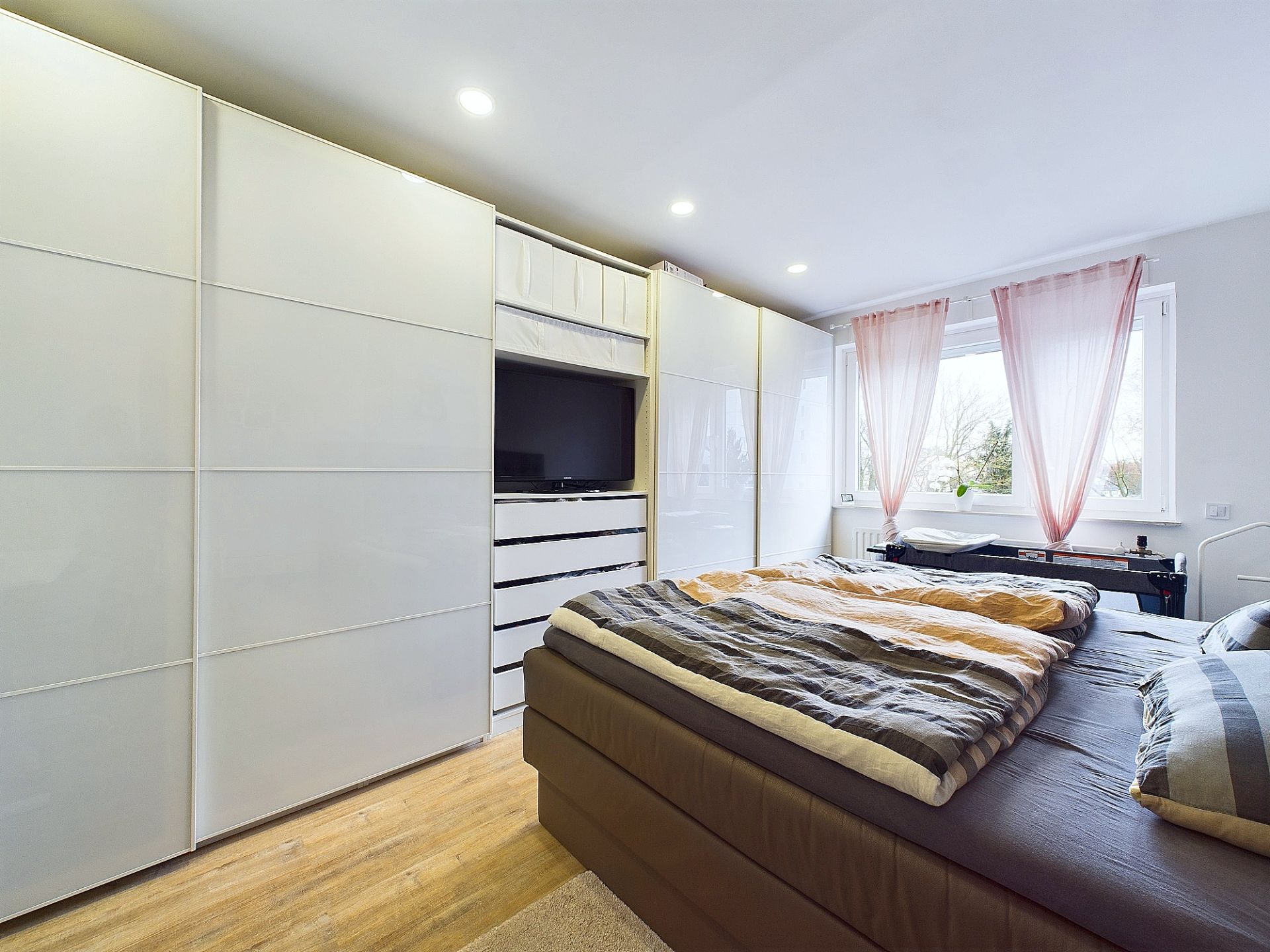 Modernes Schlafzimmer mit großem Bett, weißen Schränken und Fenstern mit rosa Vorhängen.