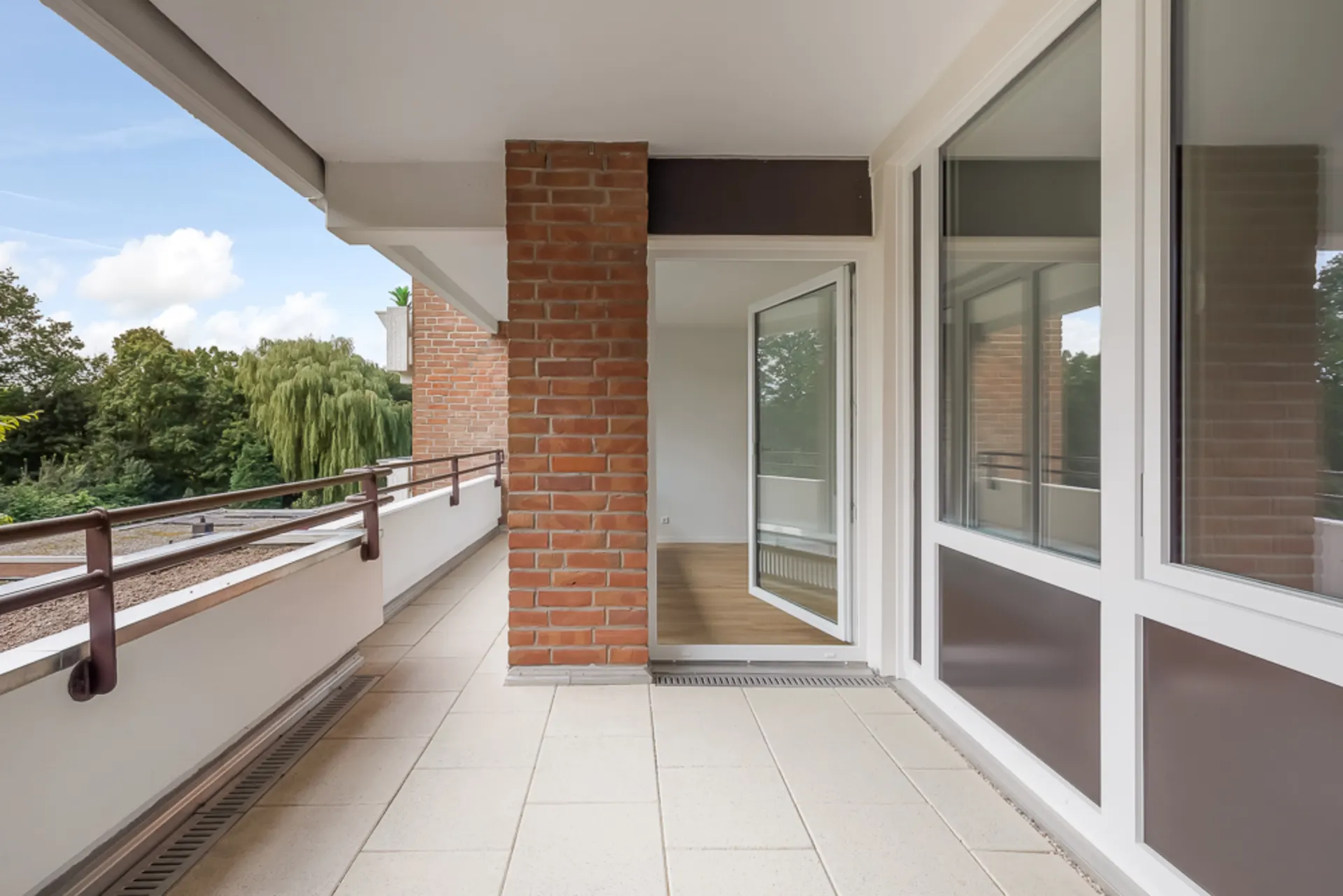Großer Balkon mit Fliesenboden, Glasgeländer und Zugang zu einem hellen Innenraum.