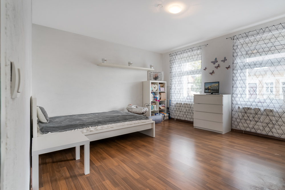 Helles Schlafzimmer mit Einzelbett, Regal, Kommode und zwei Fenstern mit Vorhängen.