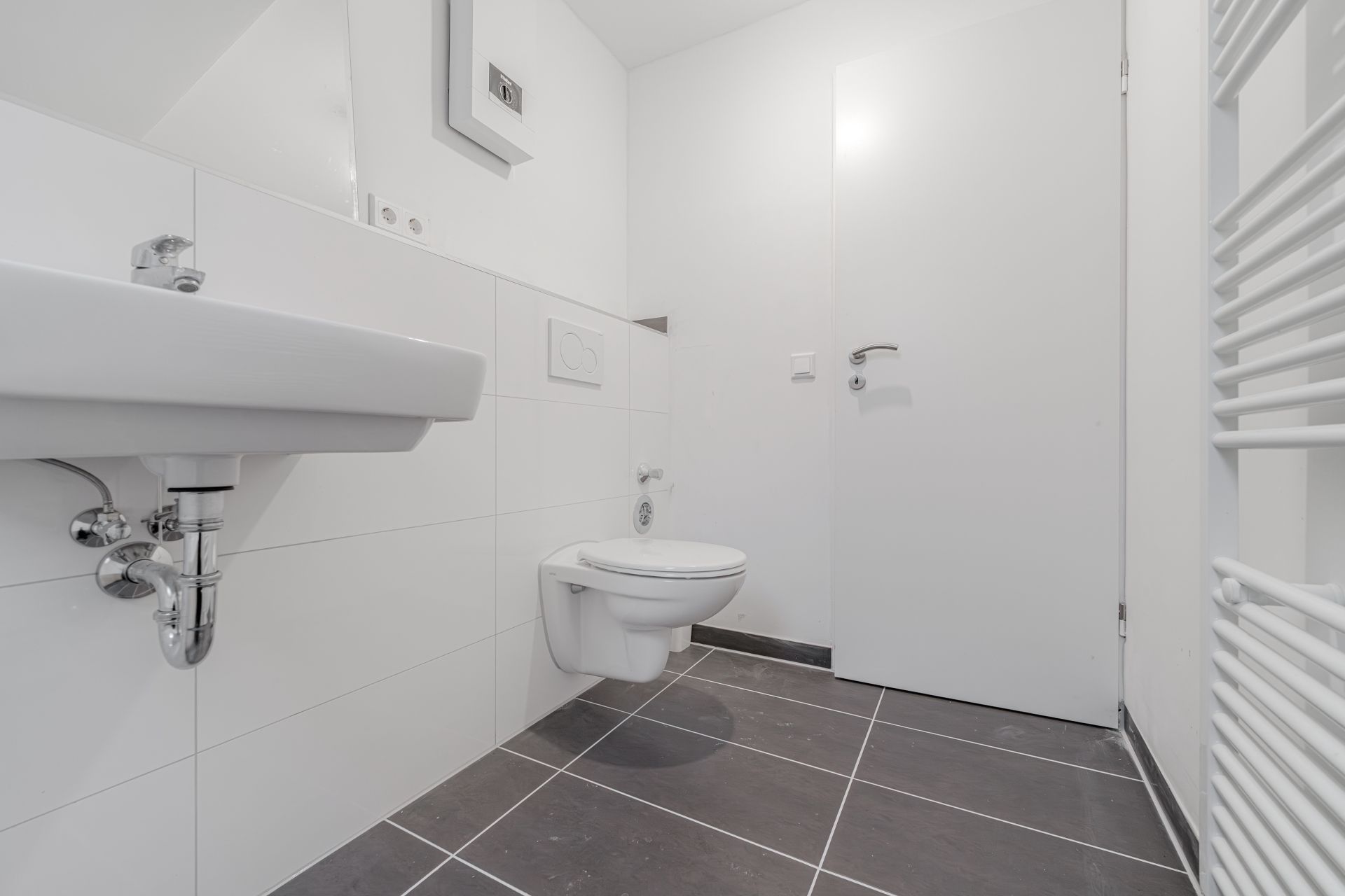 Modernes, weiß gefliestes Badezimmer mit Wand-WC, Waschbecken und dunklem Fliesenboden.