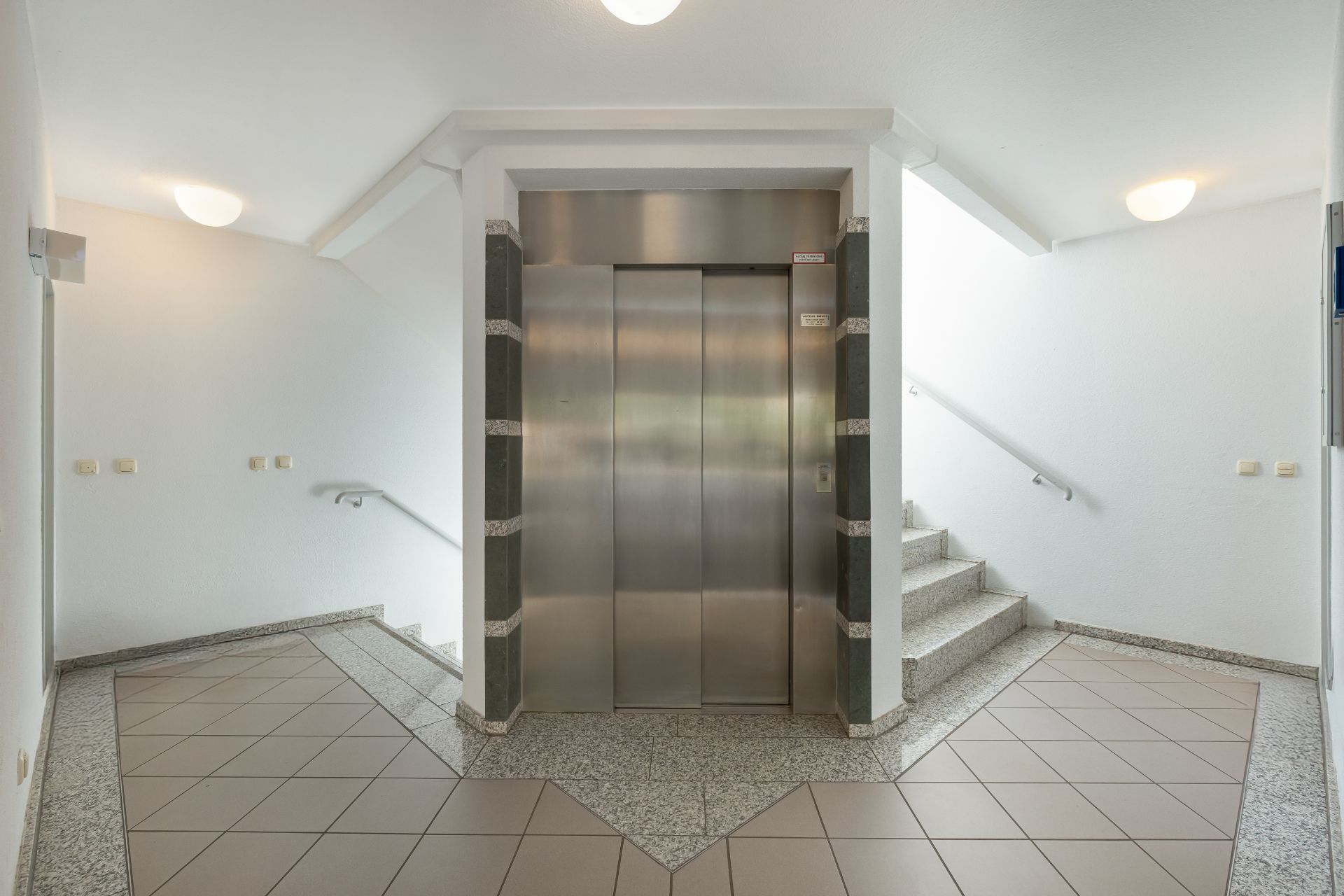 Heller Hausflur mit Aufzug in der Mitte und Treppen auf beiden Seiten.