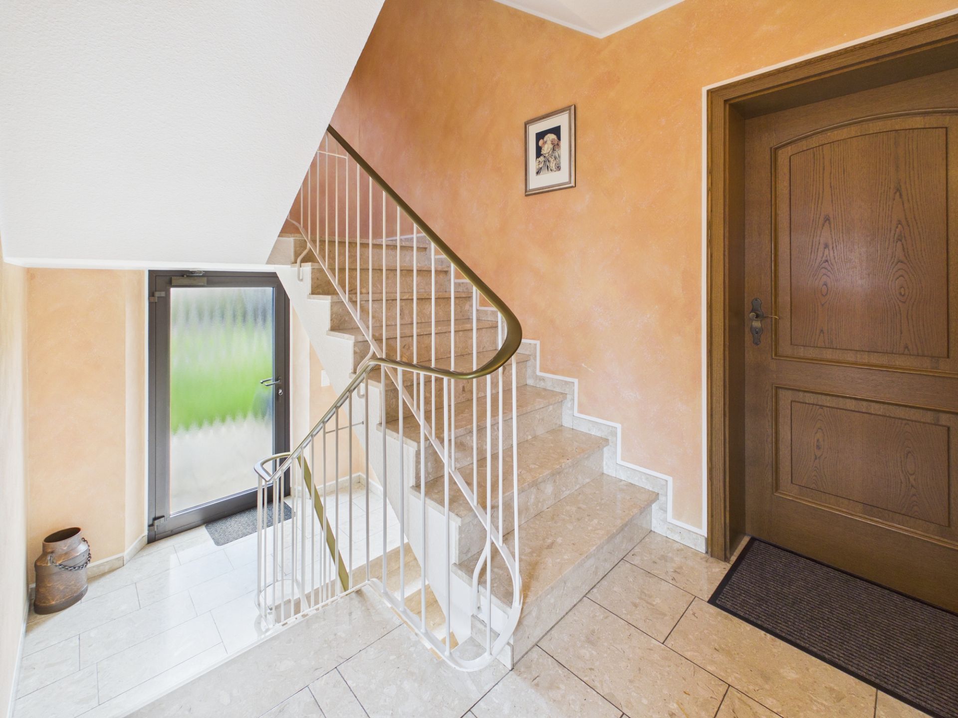 Heller Hausflur mit Steintreppe, brauner Tür und einem Bild an der Wand.