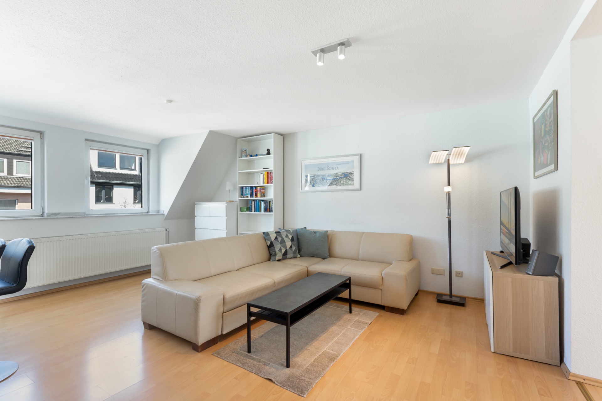 Helles Wohnzimmer mit Ecksofa, Couchtisch, Regal, Stehlampe und Fernseher auf Holzfußboden.