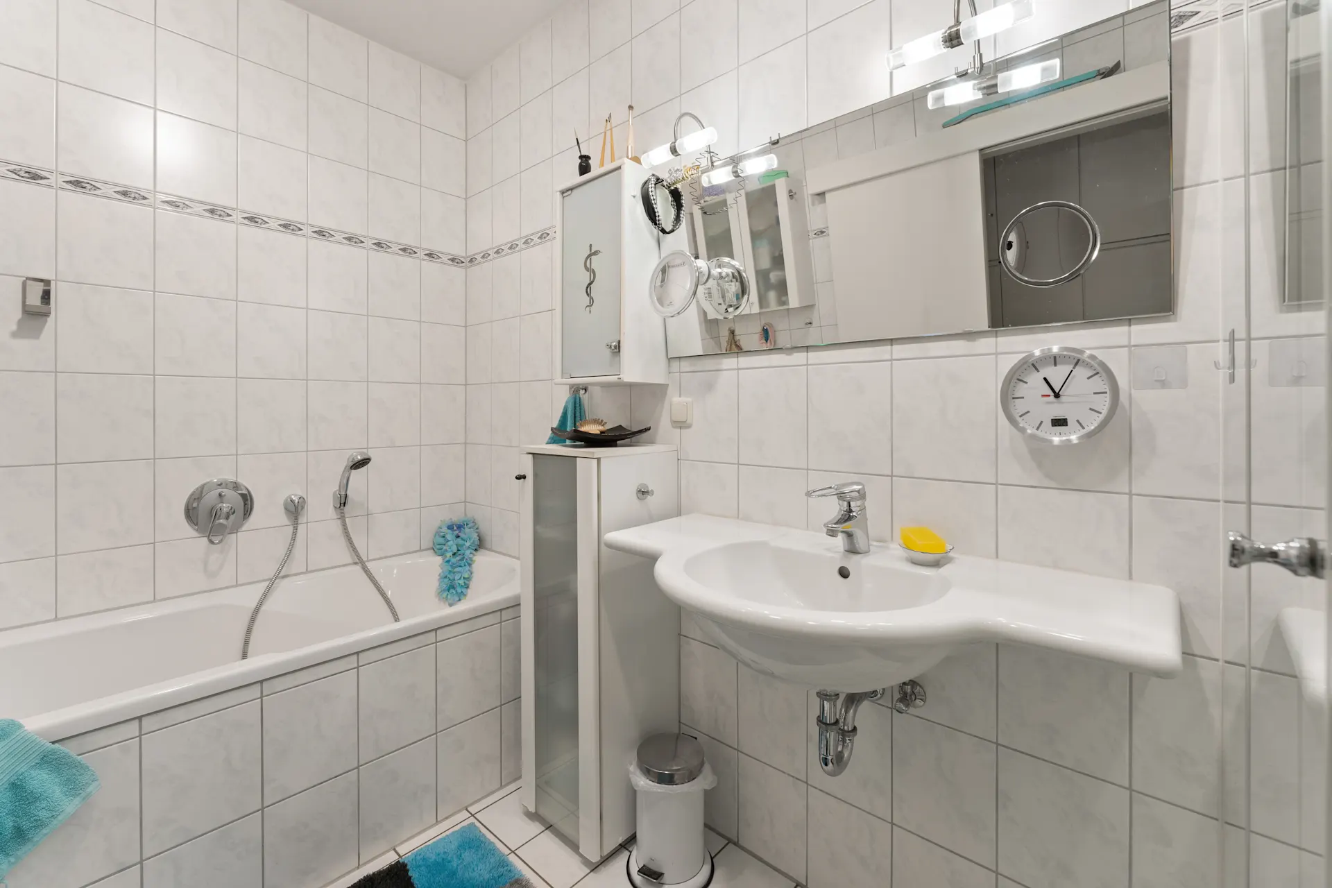 Modernes Badezimmer mit Badewanne, Waschbecken, Spiegel und Wanduhr, alles weiß gefliest.