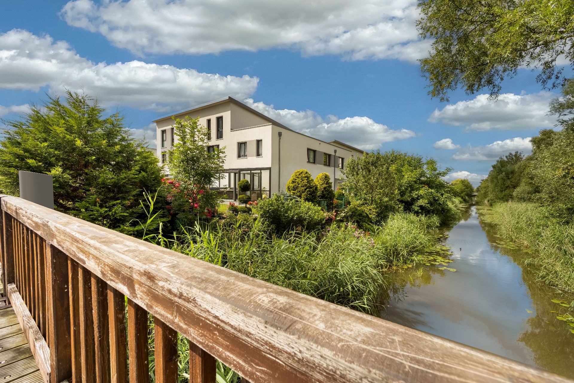 Modernes Wohnhaus mit Garten an einem kleinen Fluss, Blick von einer Holzbrücke.