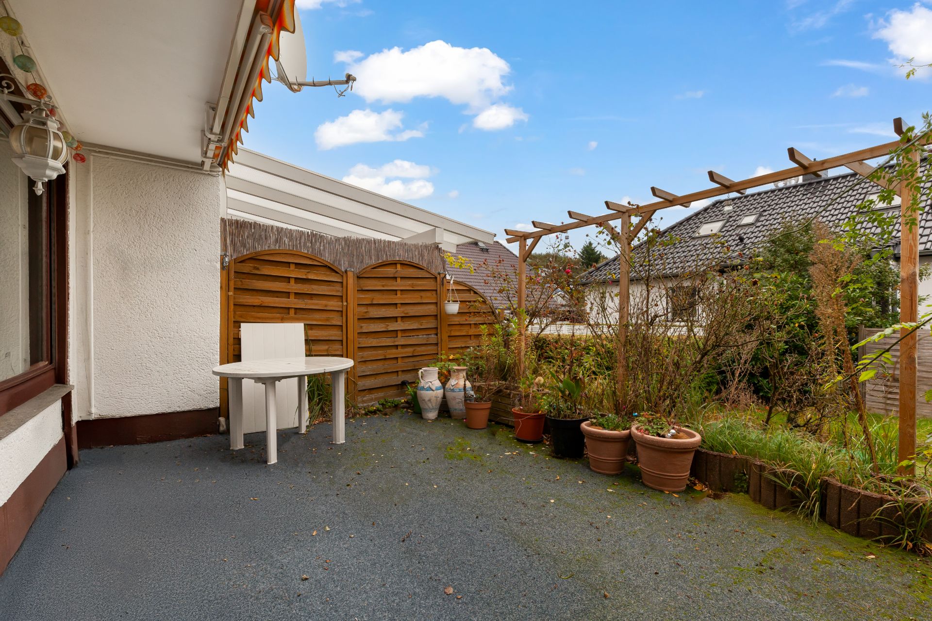 Terrasse mit kleinem runden Tisch, Stuhl und mehreren Pflanzentöpfen, daneben Holzzaun und Pergola.