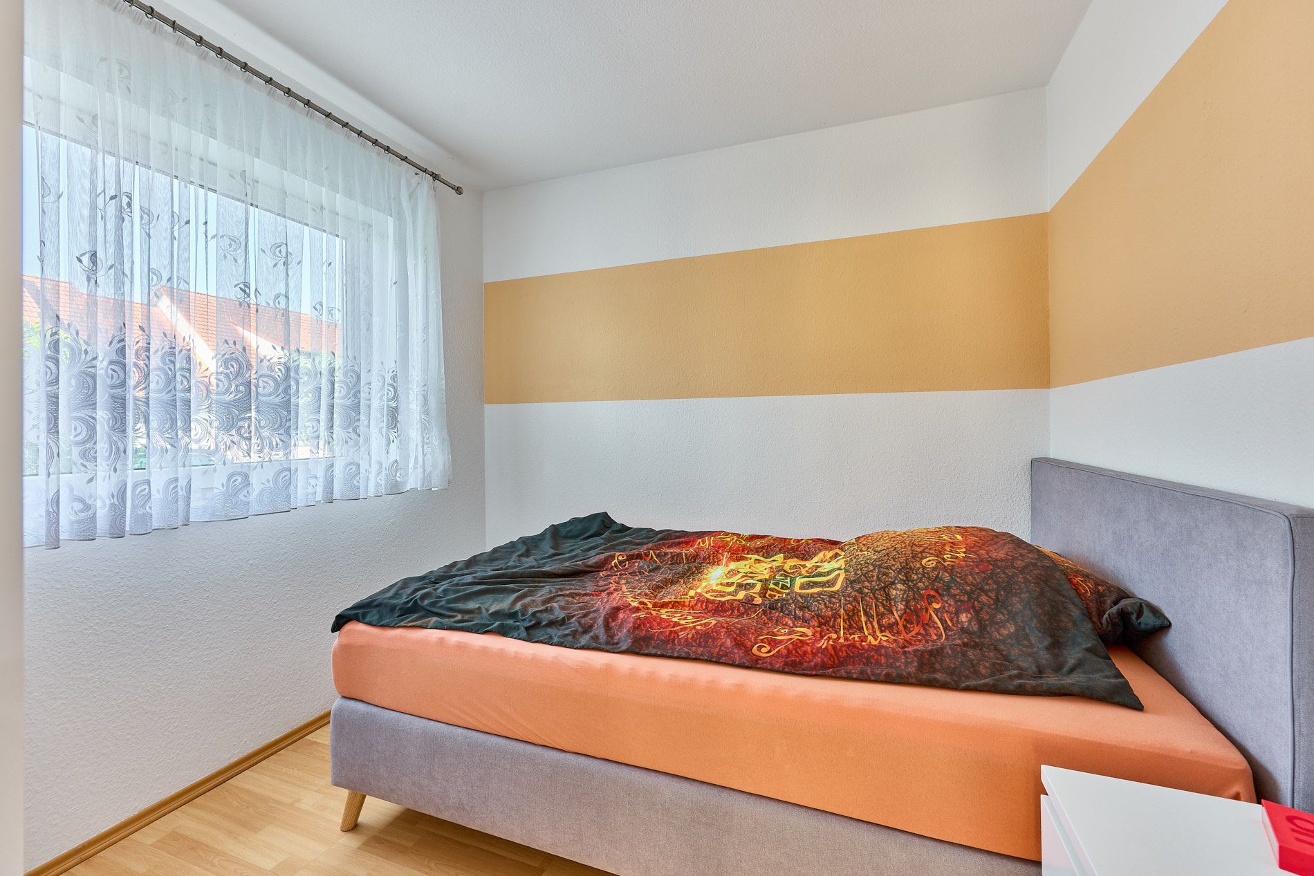 Kleines Schlafzimmer mit Einzelbett, Fenster mit Gardine und gelbem Streifen an der Wand.