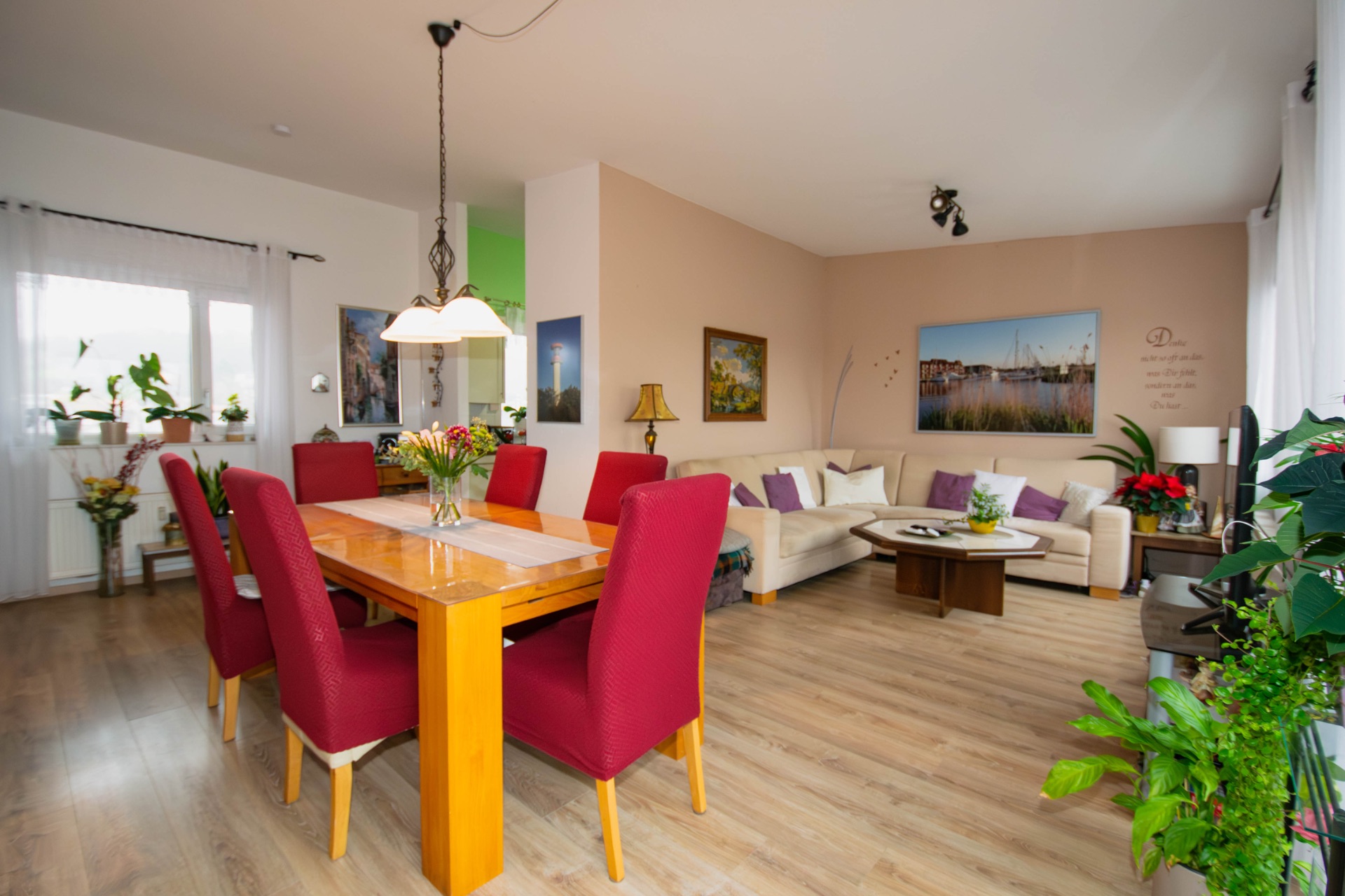 Wohnzimmer mit Esstisch, roten Stühlen, Sofa, Pflanzen und großen Fenstern.
