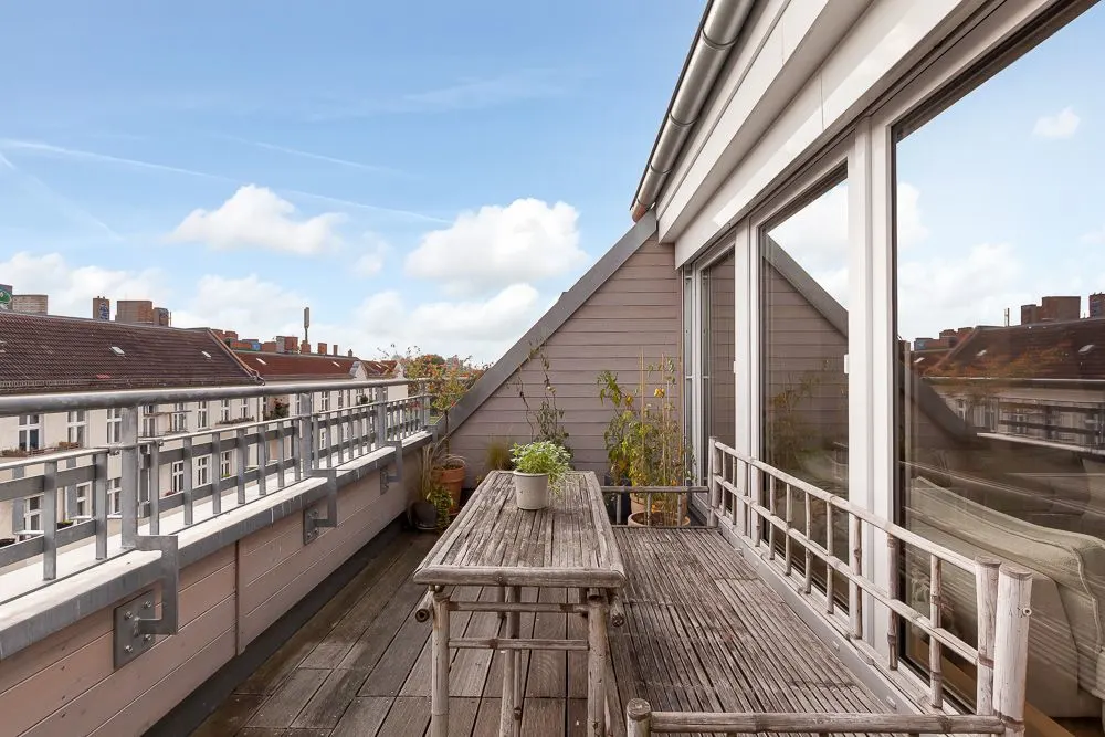 Holzbalkon mit Tisch, Stühlen und Pflanzen neben großen Fenstern einer Wohnung.