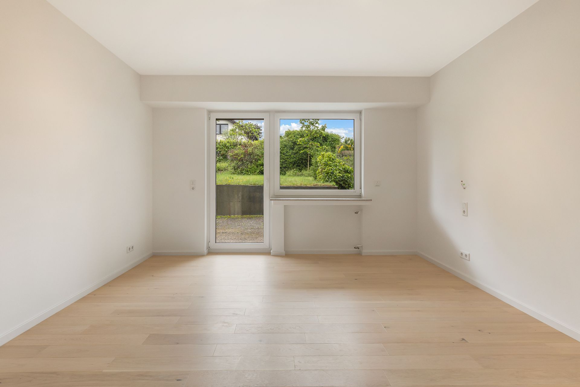 Leerer Raum mit hellem Holzboden, weiß gestrichenen Wänden und Fenster mit Blick ins Grüne.