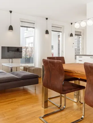 Helles Wohnzimmer mit grauem Sofa, Esstisch und braunen Stühlen auf Parkettboden.