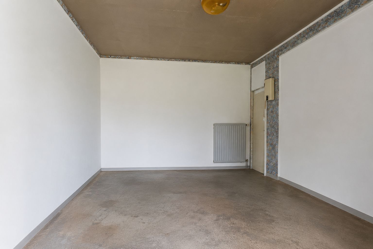 Leerer Raum mit weiß gestrichenen Wänden, Heizkörper und braunem Boden.