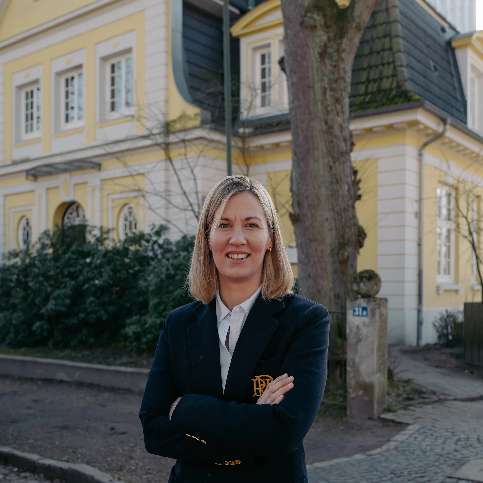 Frau steht mit verschränkten Armen vor einem gelben Wohnhaus mit weißen Fenstern.
