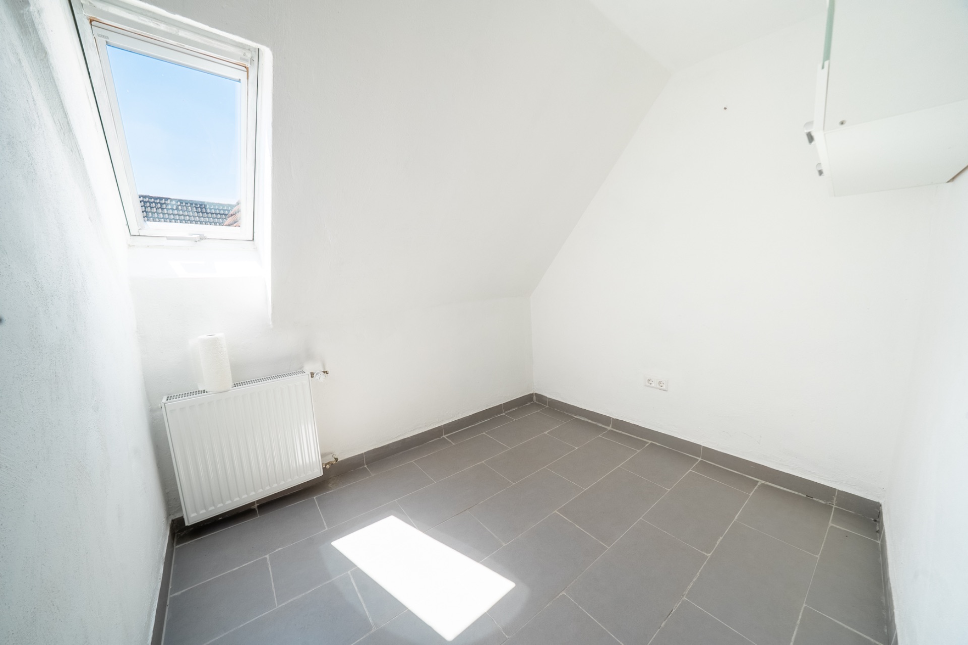 Kleiner, leerer Raum mit grauen Fliesen, weißer Wand, Fenster und Heizkörper.