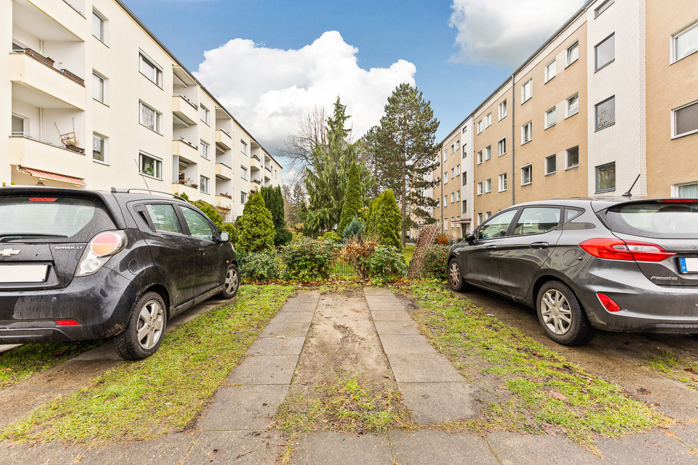 Zwei Autos parken zwischen Mehrfamilienhäusern, im Hintergrund grüne Büsche und Bäume.