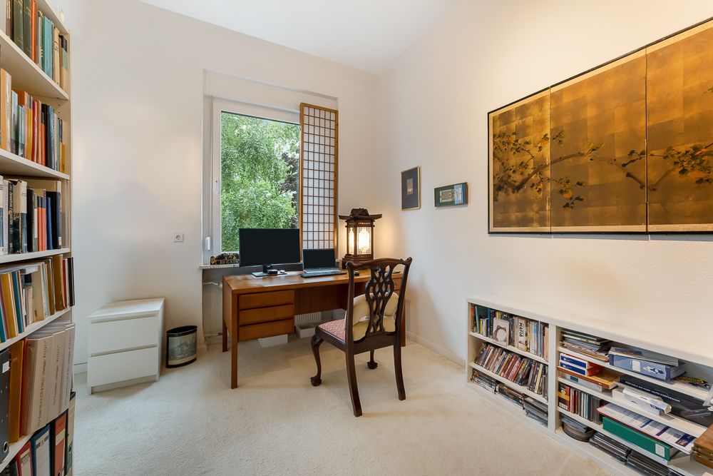 Helles Arbeitszimmer mit Schreibtisch, Stuhl, Bücherregalen und Computer am Fenster.