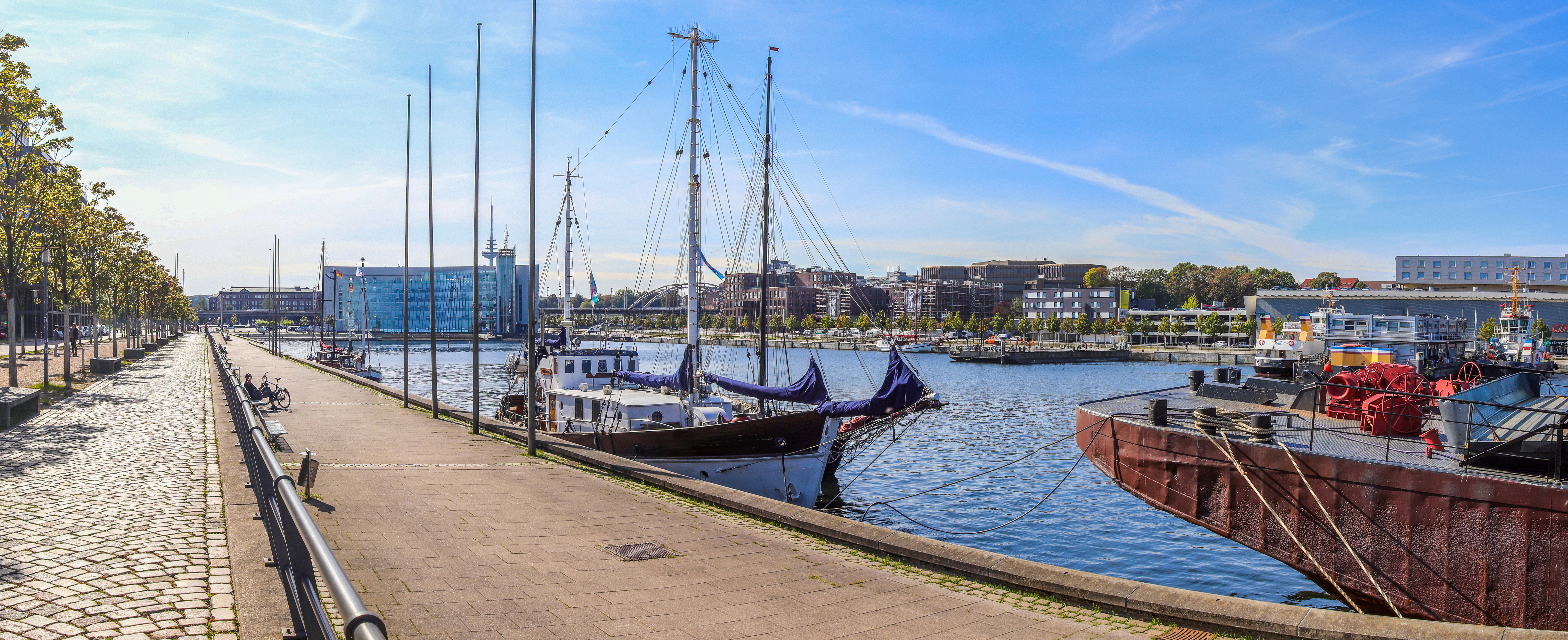 Promenade mit Kopfsteinpflaster am Wasser, Segelboote und Schiffe im Hafen.