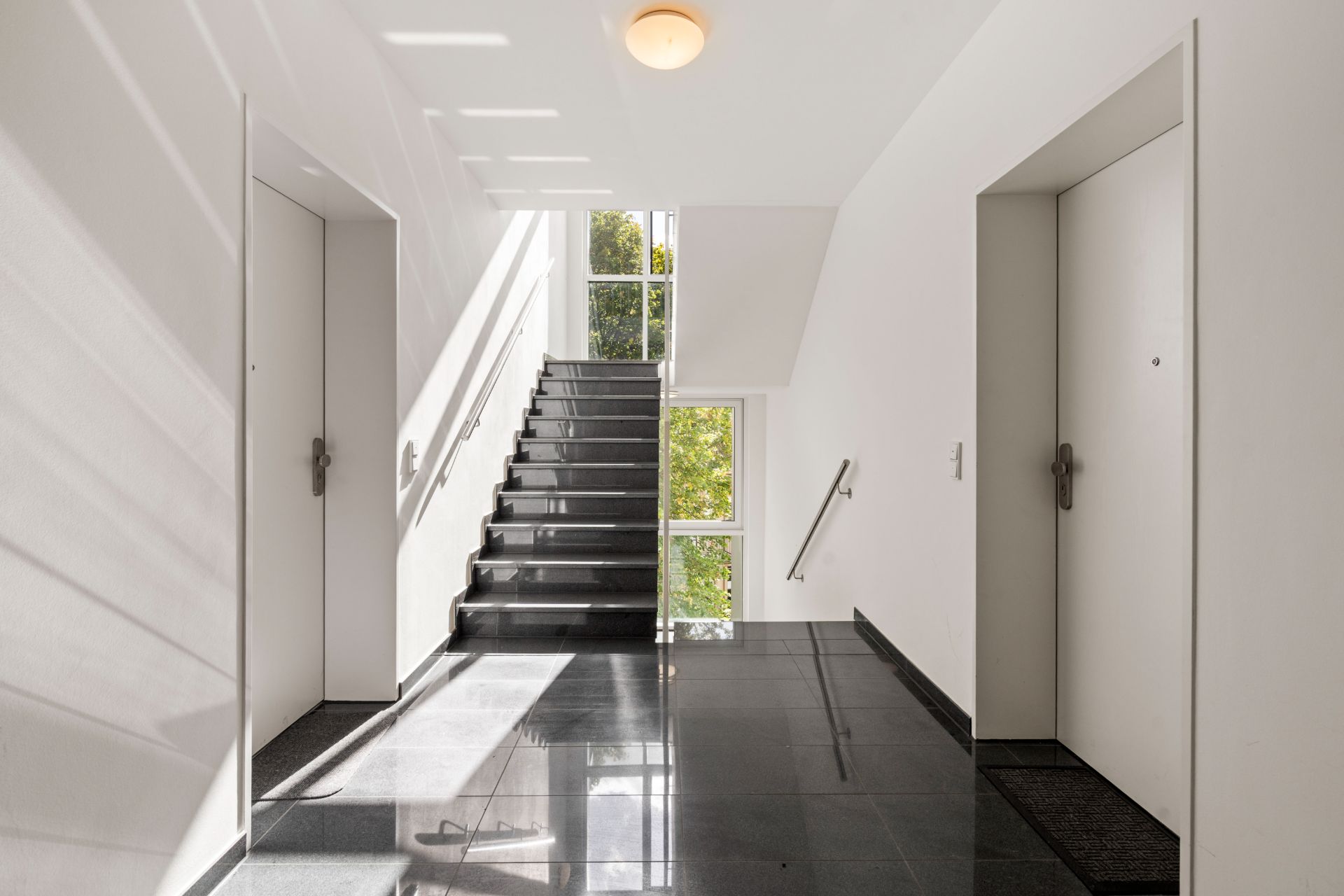 Heller Hausflur mit zwei Wohnungstüren und einer Treppe vor großen Fenstern.