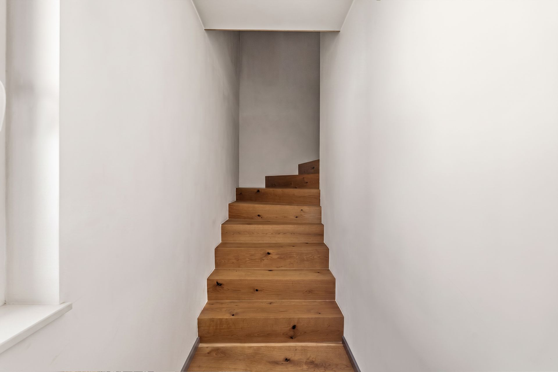 Eine schmale Holztreppe in einem weißen Flur, die nach oben führt.