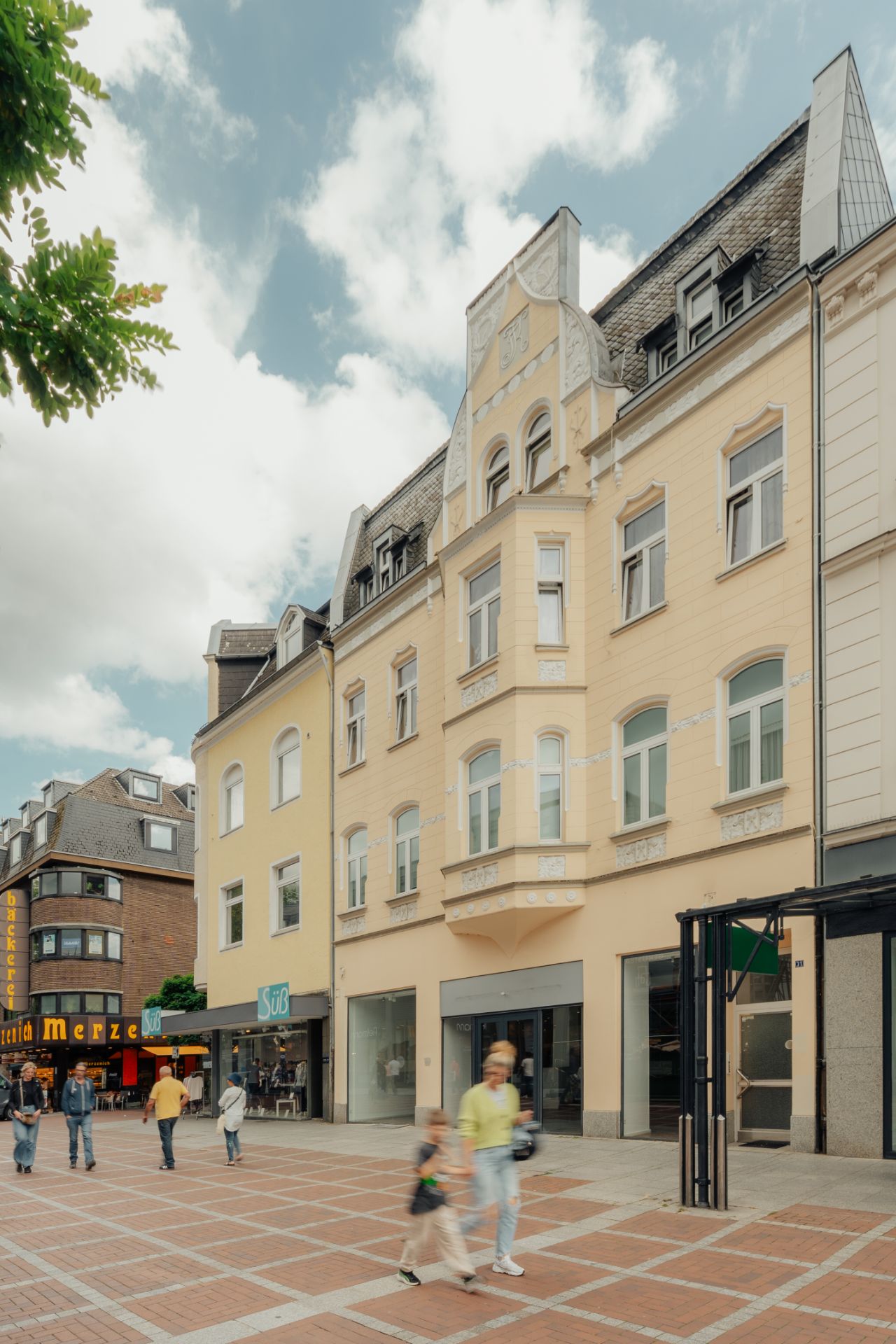 Mehrstöckiges Wohnhaus mit heller Fassade und Geschäften im Erdgeschoss, Passanten auf dem Gehweg.
