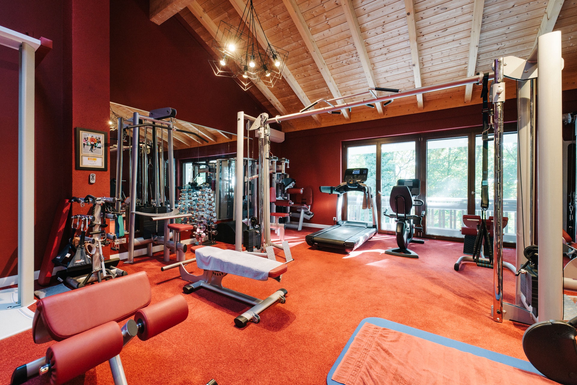Fitnessraum mit roten Wänden und Teppich, mehreren Fitnessgeräten und großen Fenstern.