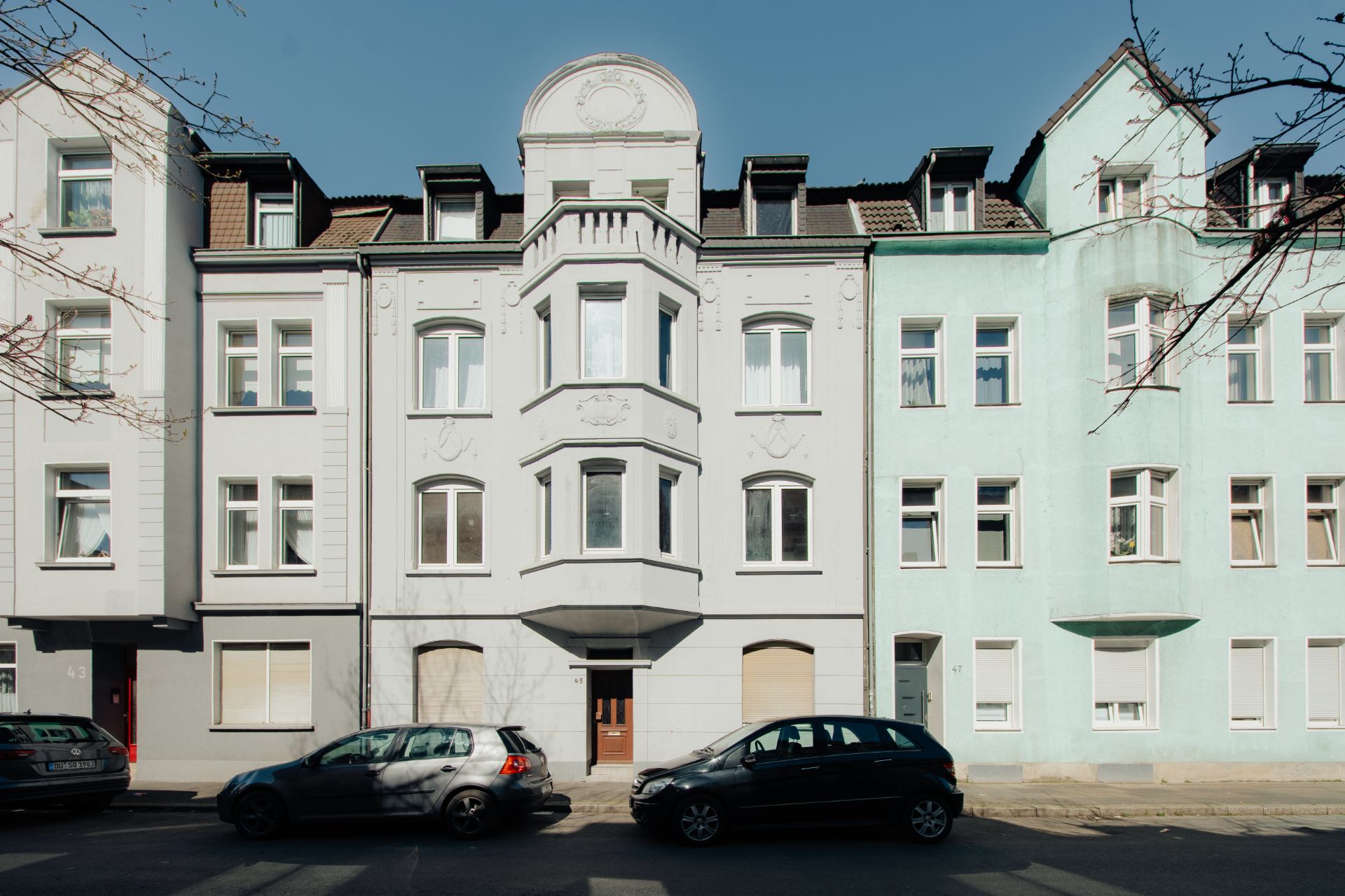 Mehrfamilienhaus mit Erker und mehreren Fenstern, zwei Autos parken davor auf der Straße.