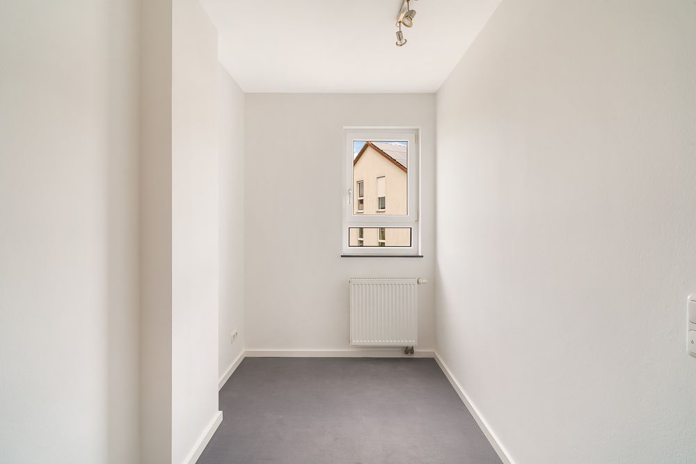 Kleiner leerer Raum mit grauem Boden, weißem Heizkörper und Fenster an der Stirnwand.