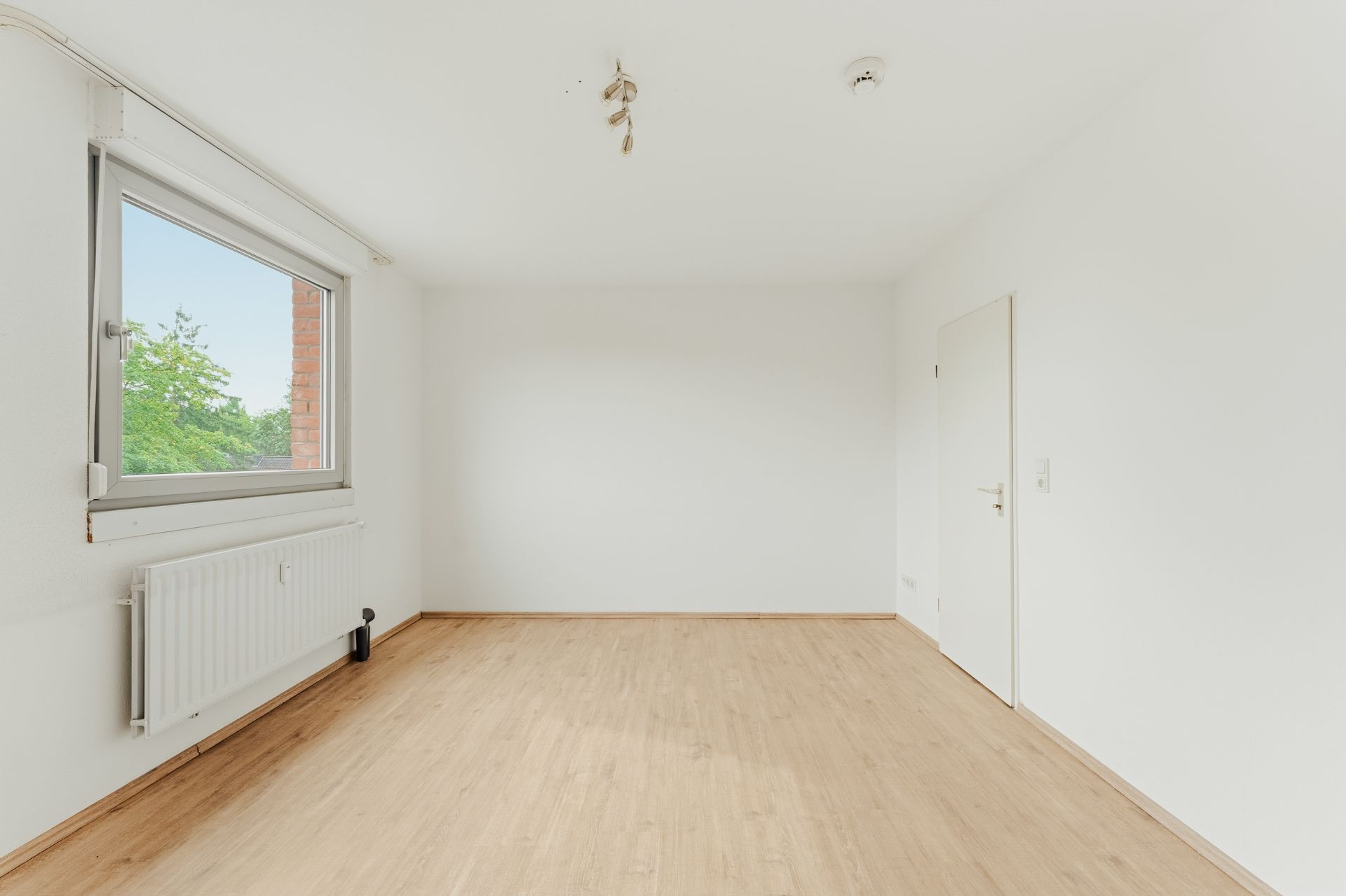 Helles, leeres Zimmer mit Holzboden, weißer Wand, Fenster und Heizung.