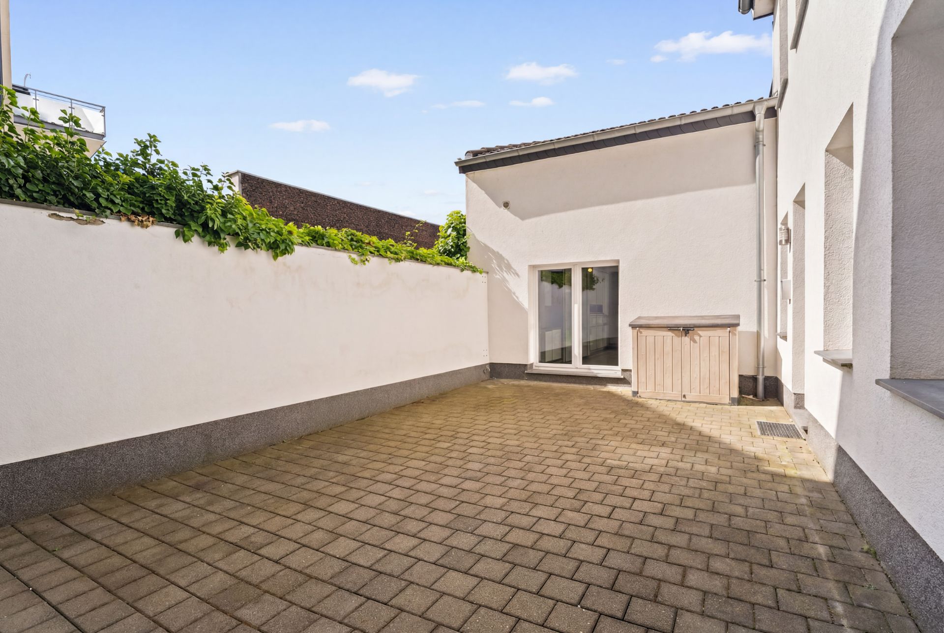 Gepflasterter Innenhof mit weißer Wand, Kletterpflanzen und Zugangstür.