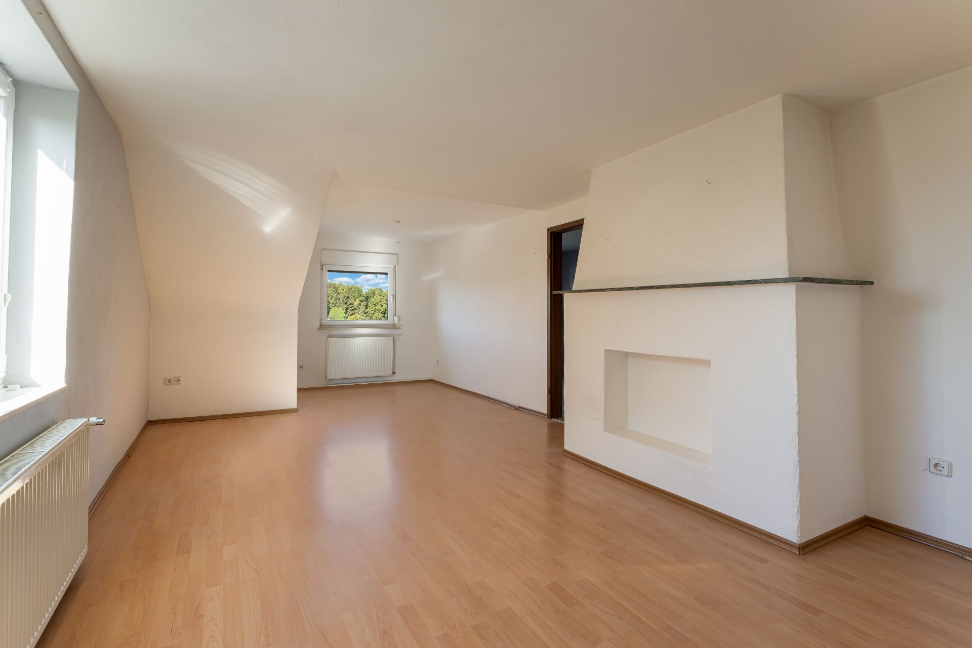 Heller, leerer Wohnraum mit Holzfußboden, weiß gestrichenen Wänden und einem Fenster am Ende des Raumes.