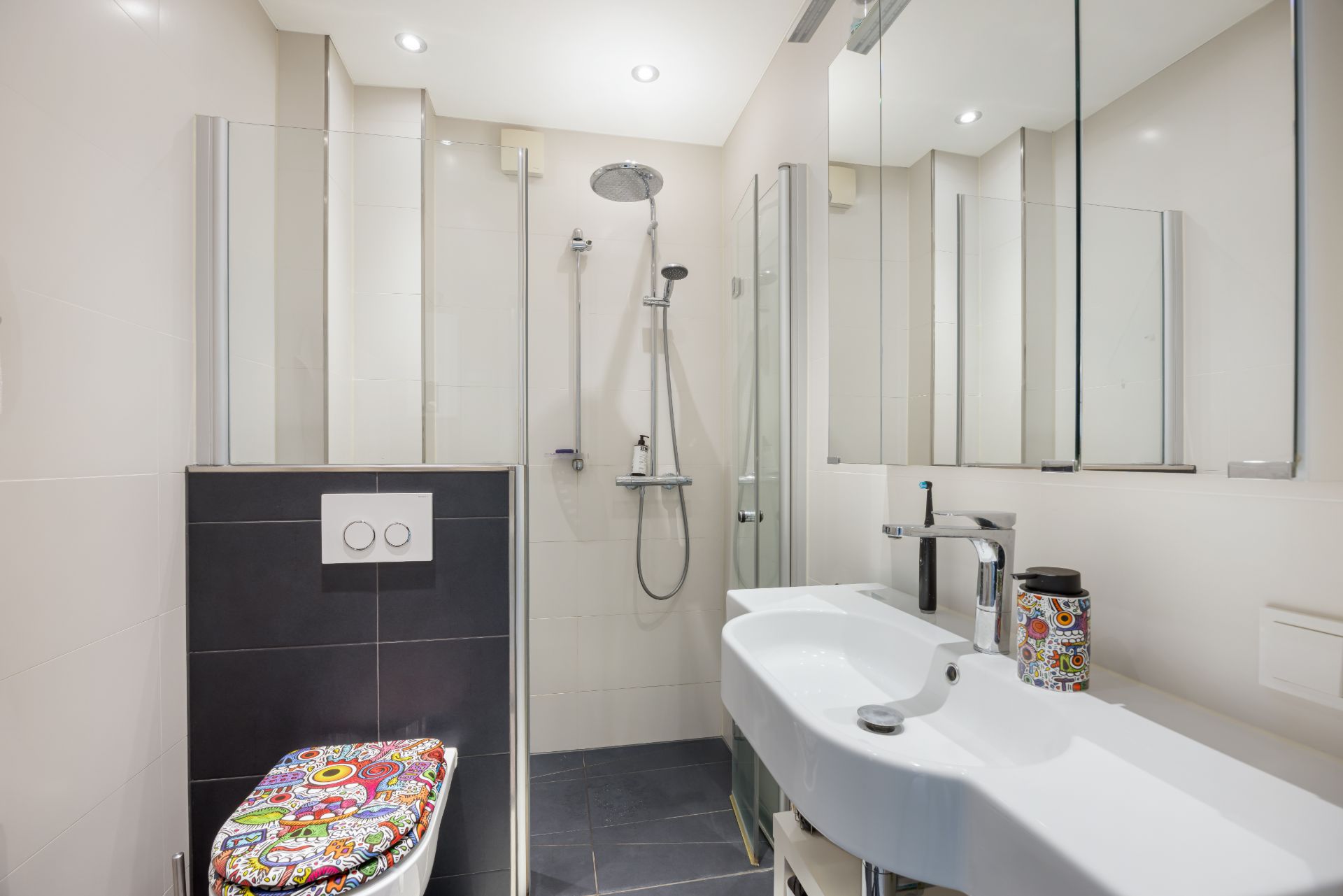 Modernes Bad mit Dusche, Waschbecken, Spiegel und WC mit buntem Toilettendeckel.