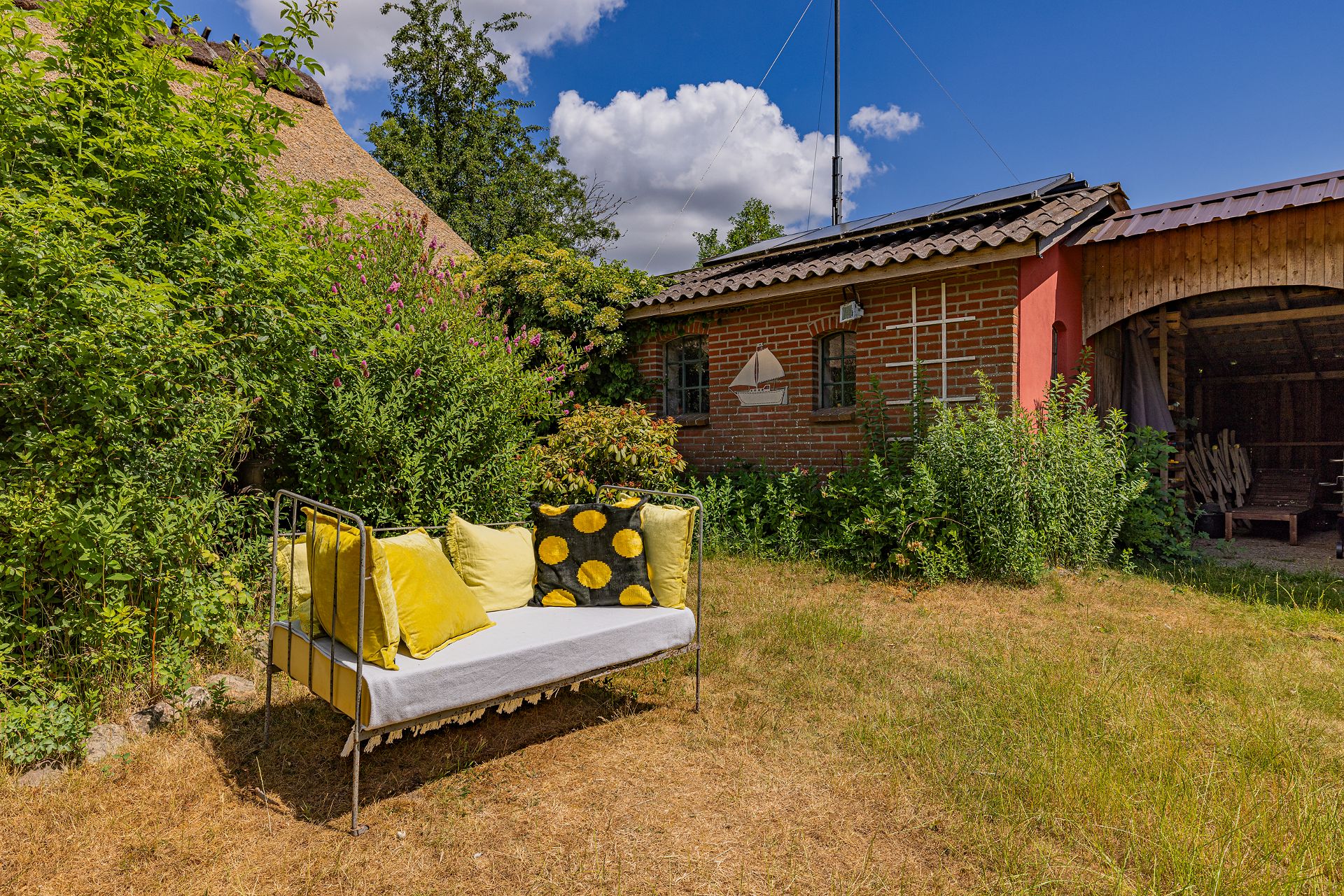 Garten mit Metallsofa und gelben Kissen vor einem Backsteinhaus an einem sonnigen Tag.