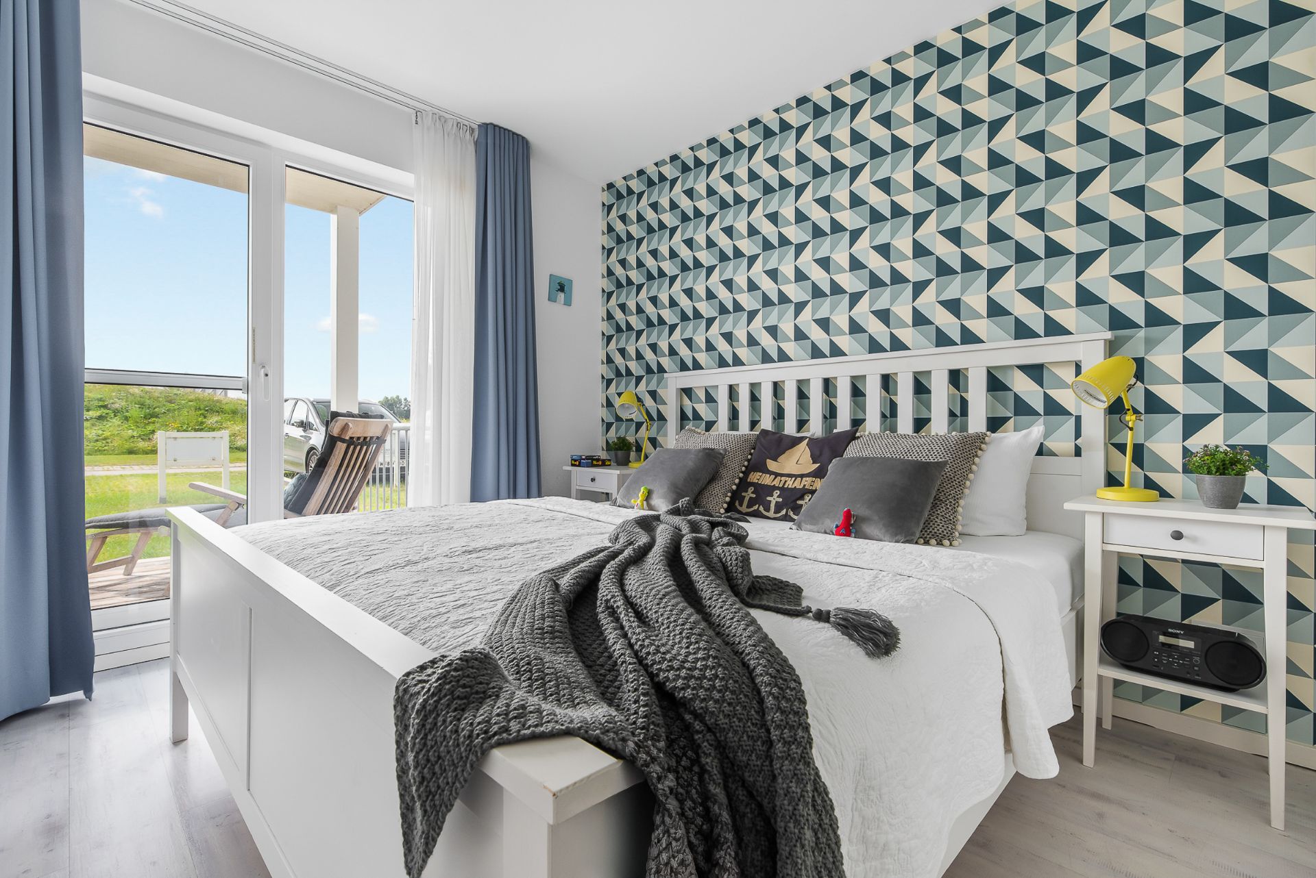 Helles Schlafzimmer mit gemusterter Akzentwand, Doppelbett und Zugang zu einem Balkon.