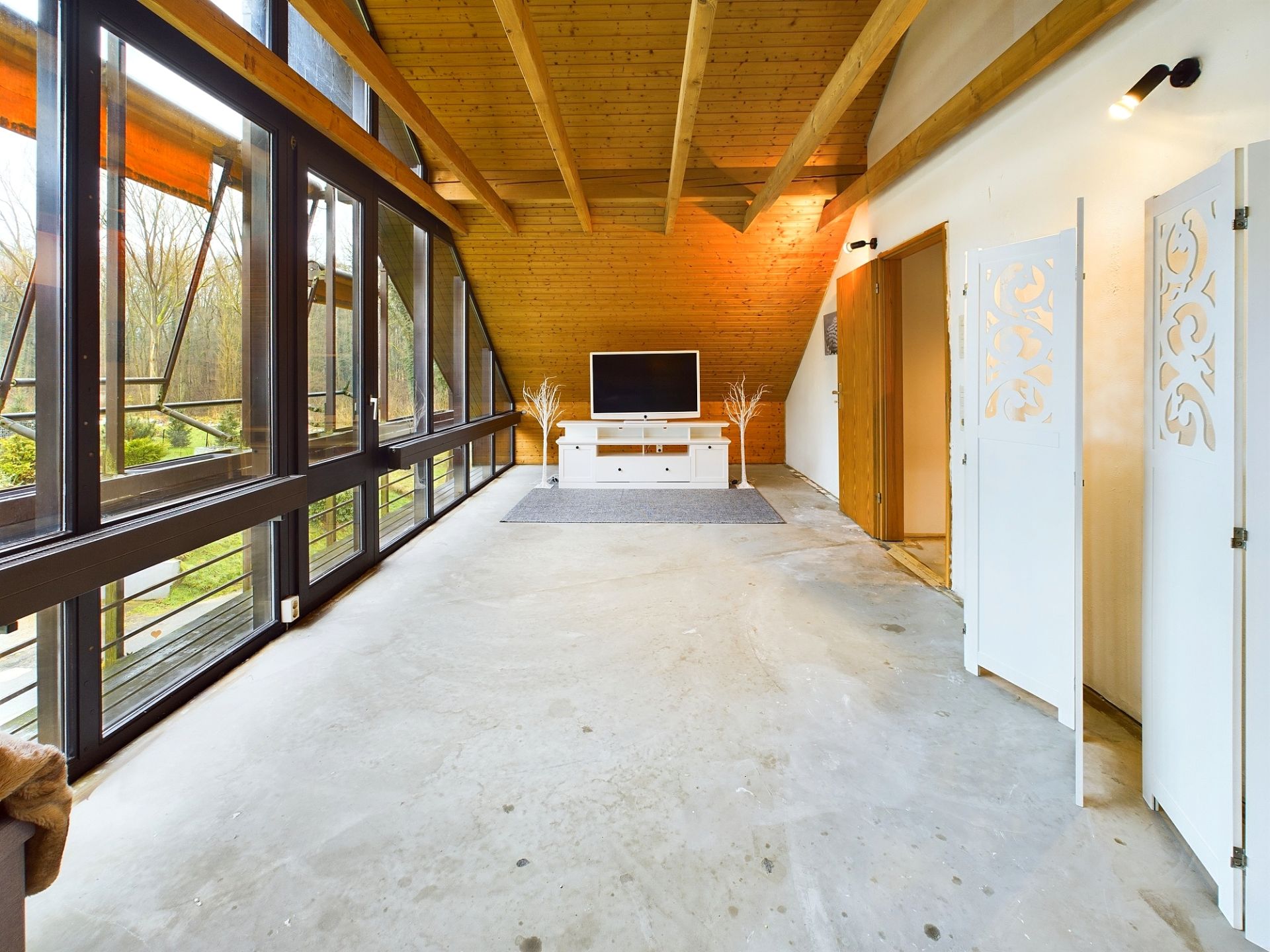 Wohnzimmer mit großen Fenstern, Holzdach, Fernseher und weißem TV-Schrank.
