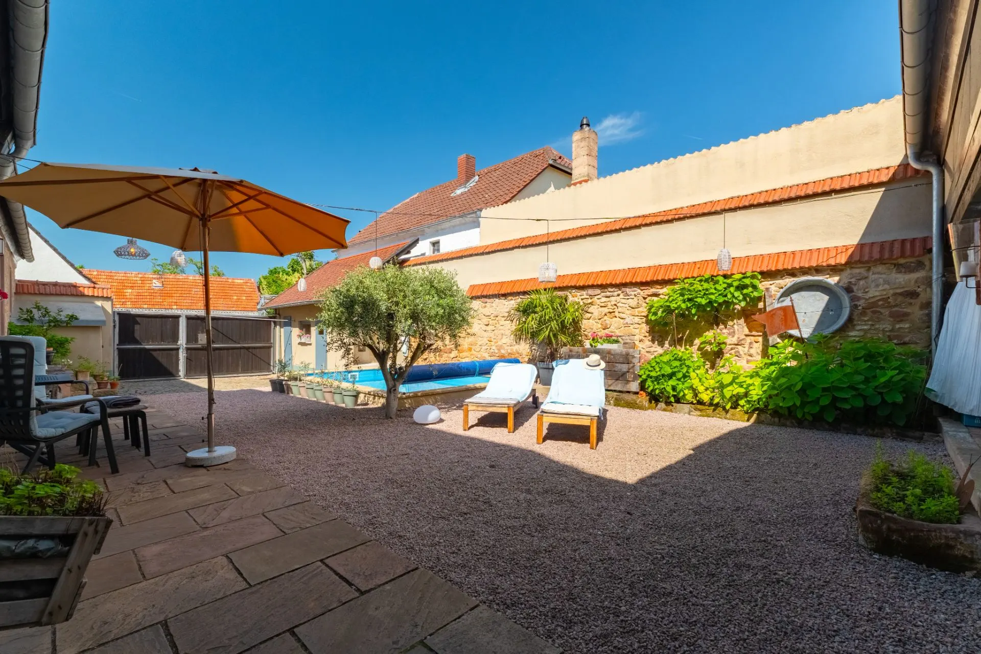 Hof mit zwei Liegestühlen, Sonnenschirm, kleinem Pool und bepflanzten Beeten an einer Steinmauer.