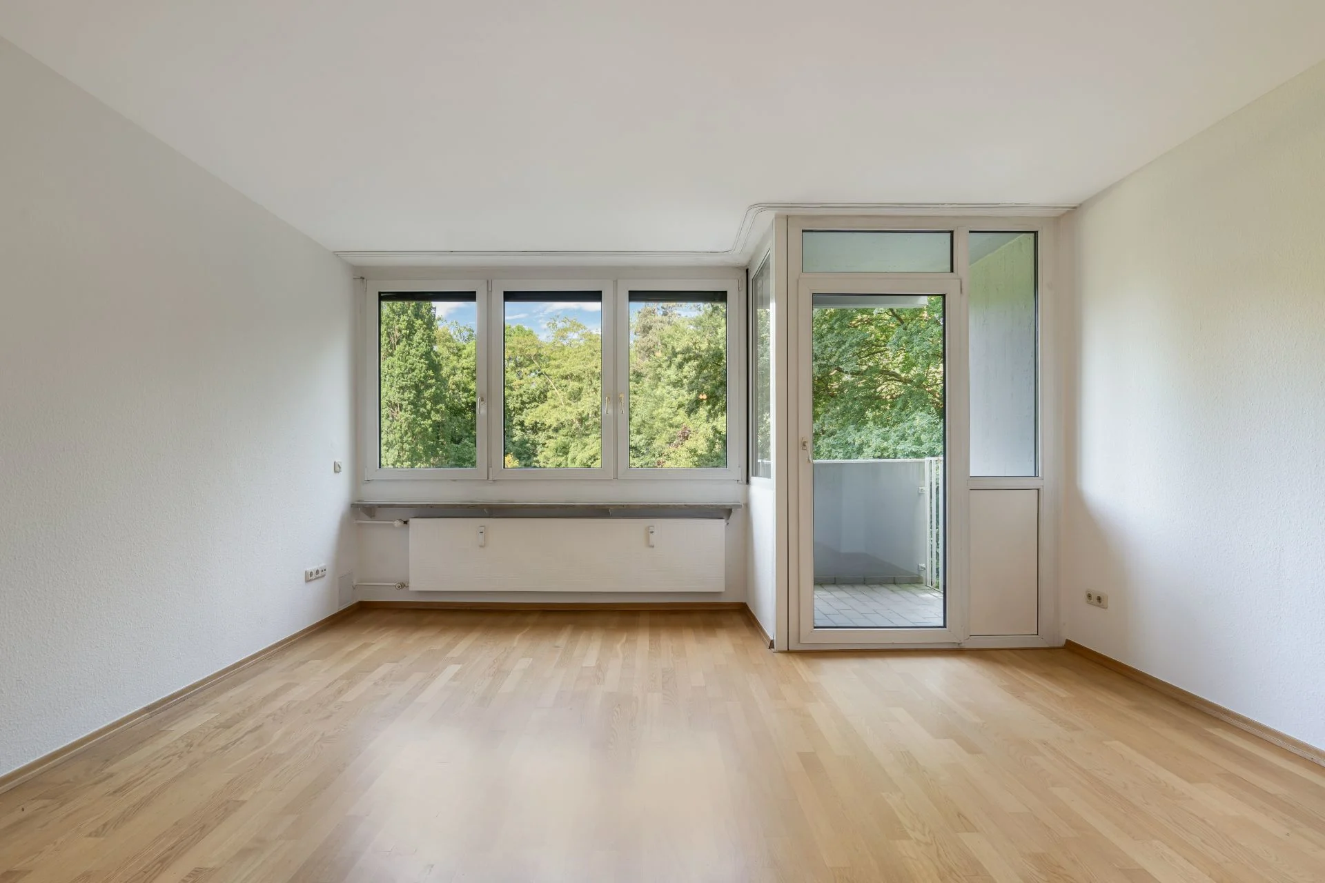 Leeres Zimmer mit Holzfußboden, großen Fenstern und Tür zu einem Balkon mit Blick ins Grüne.