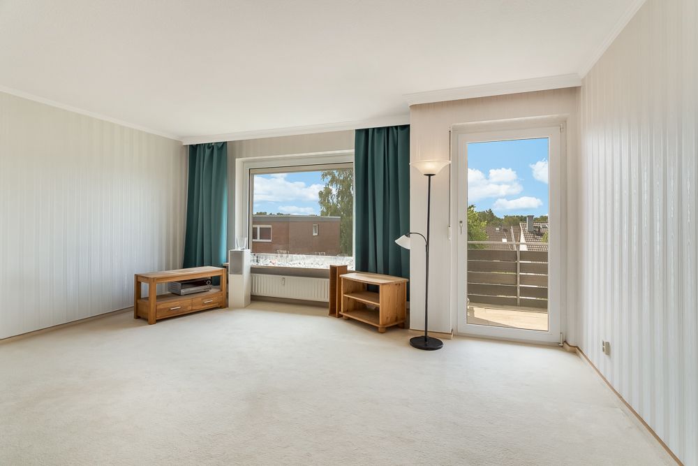 Helles Wohnzimmer mit Teppich, zwei Holzmöbeln, Stehlampe, Fenster und Tür zum Balkon.